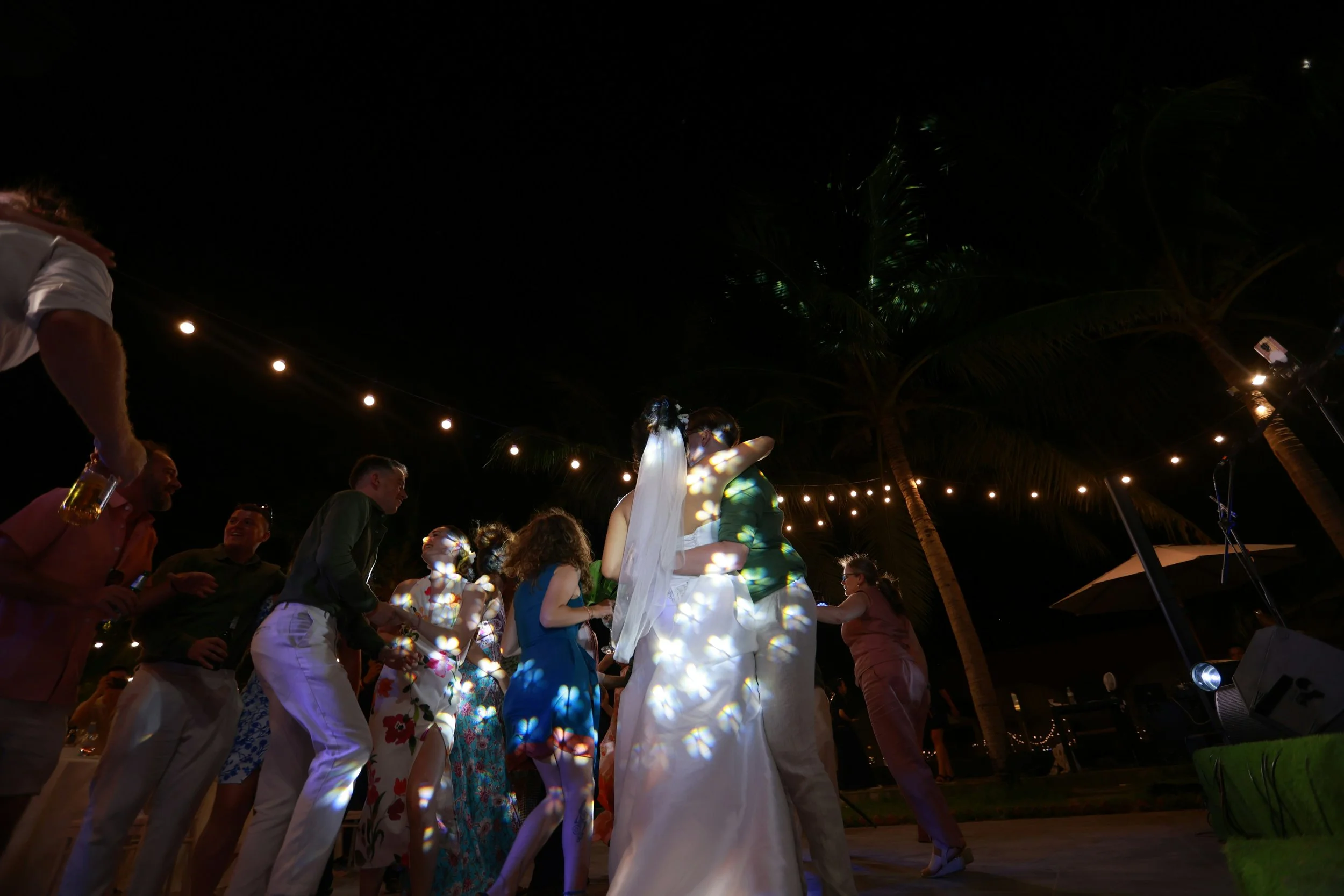 Couple dancing and hugging at a nighttime outdoor wedding reception with string lights and palm trees, surrounded by guests.