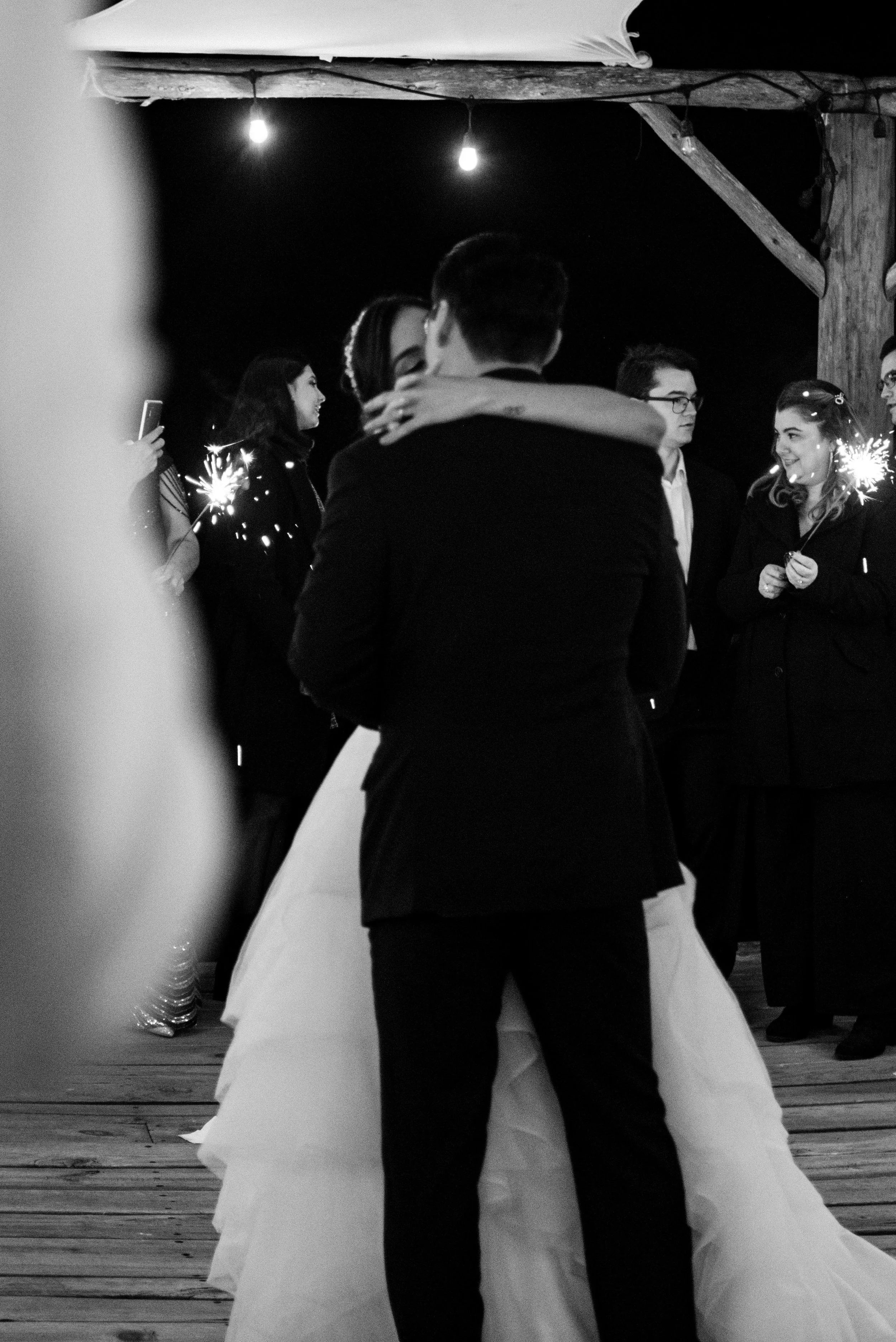 A couple in wedding attire sharing a kiss during their wedding reception at night, with guests holding sparklers in the background.