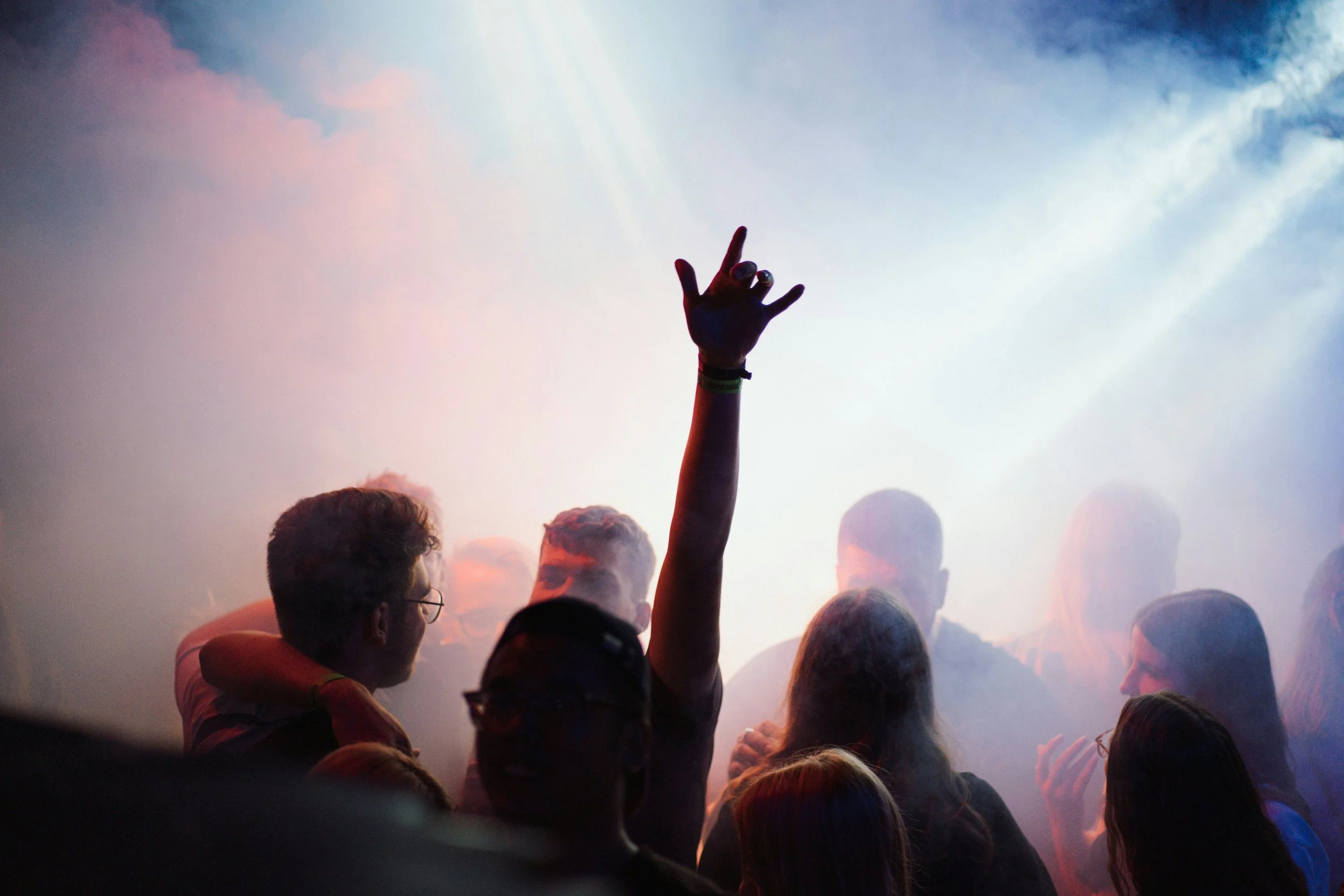 People dancing and socializing at a club or concert with colorful lighting and smoke, one person's arm raised in the air.