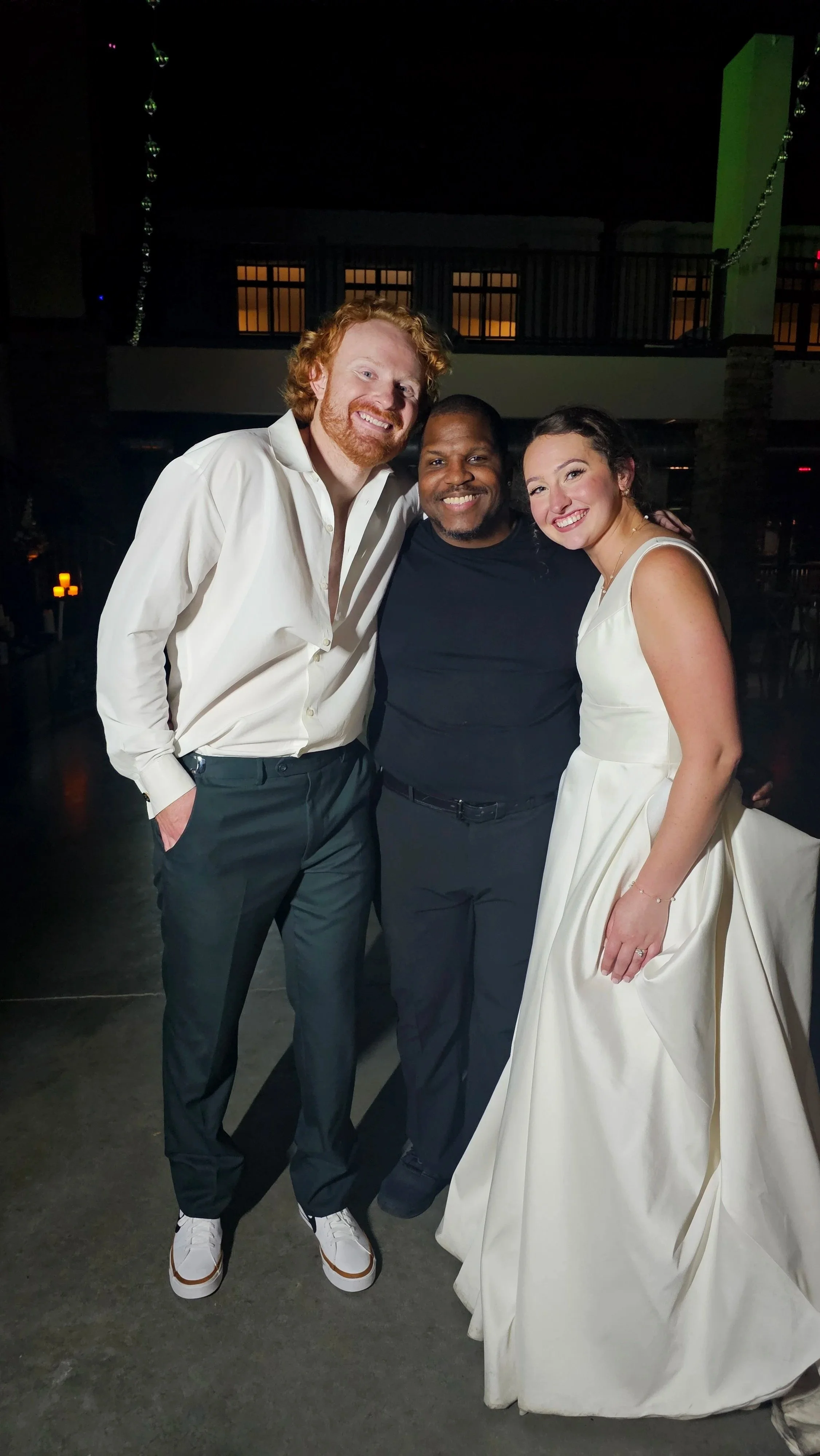 Three people standing together, smiling, at a social event or celebration. The person on the left has red hair and a beard, wearing a white shirt and dark trousers. The person in the middle has dark skin, short hair, and is wearing a black shirt. The person on the right has long hair and is wearing a white gown, possibly a wedding dress.
