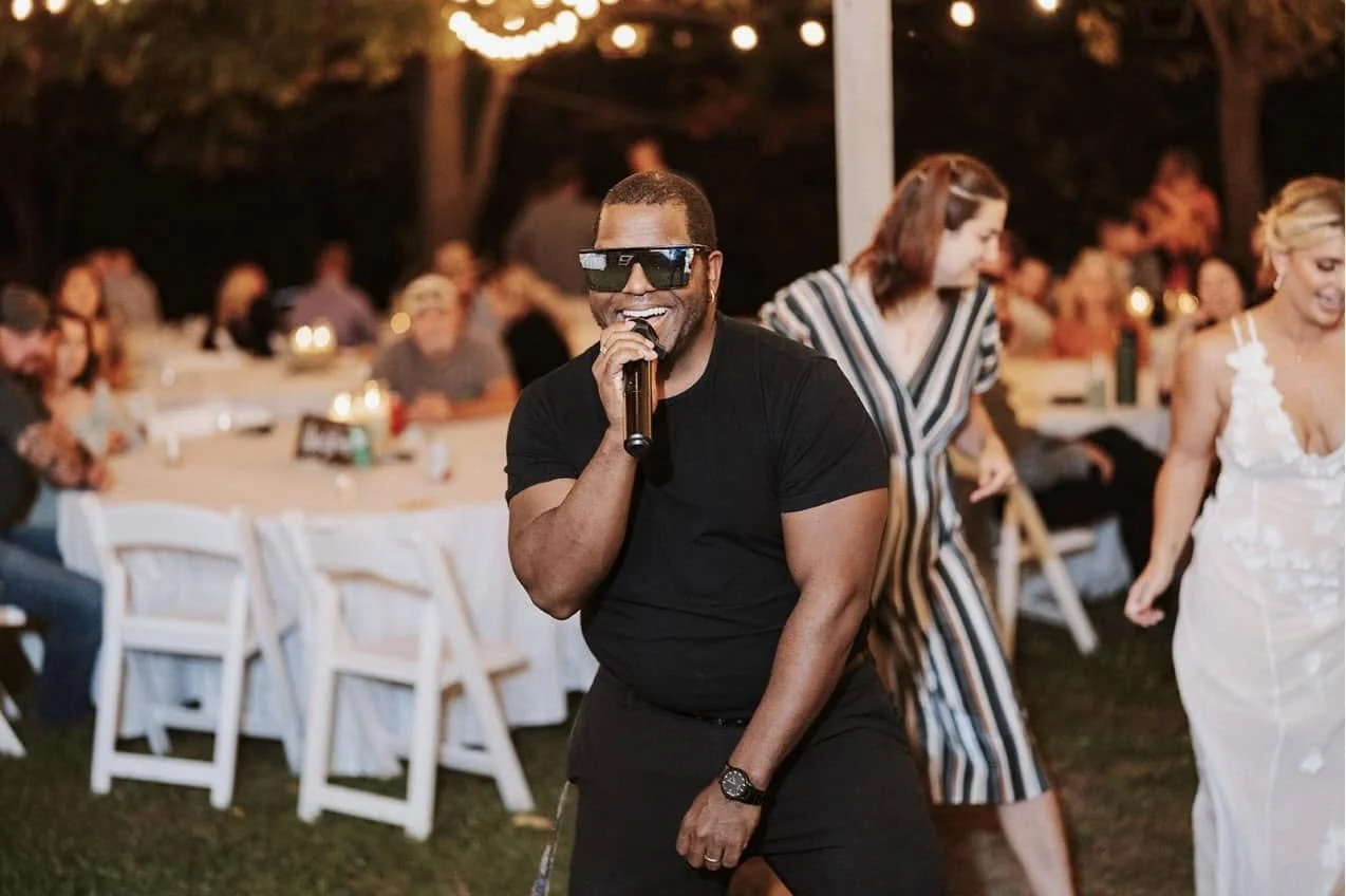A man wearing sunglasses and a black t-shirt holding a microphone at an outdoor event, with people sitting at tables and string lights in the background.