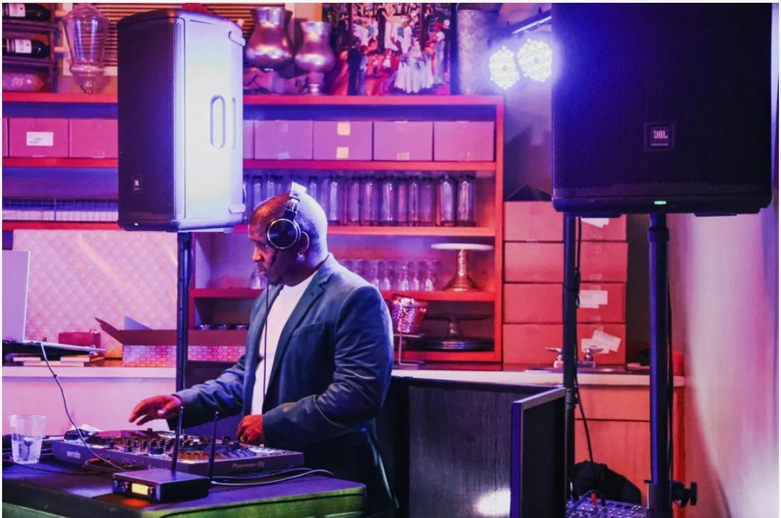 A man wearing a blazer and headphones DJing with equipment in a room with colorful lighting and shelves of glassware and books.