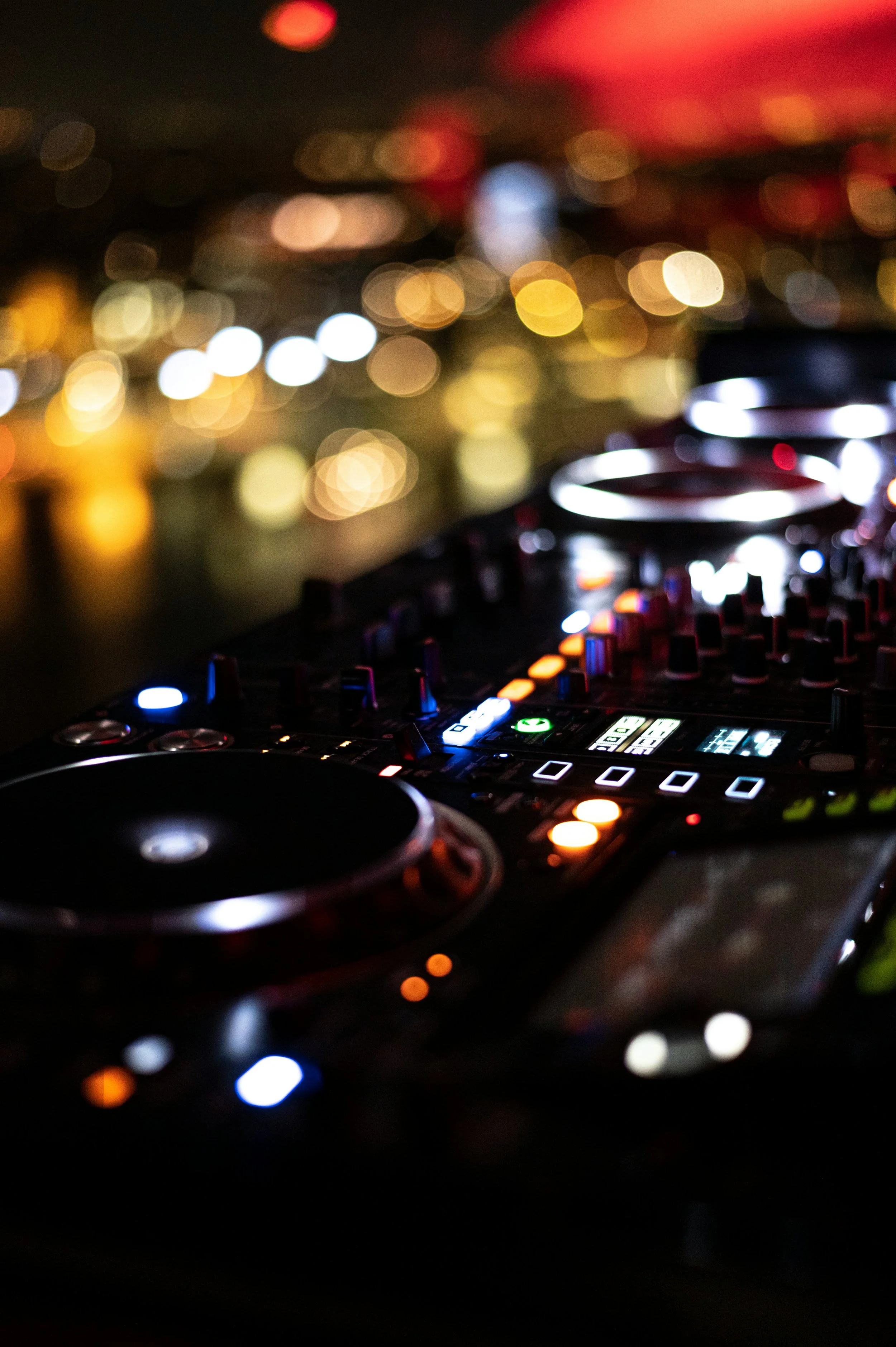 A DJ mixing music on a professional DJ controller with colorful LED lights, blurred city lights in the background, and a dark, vibrant atmosphere.