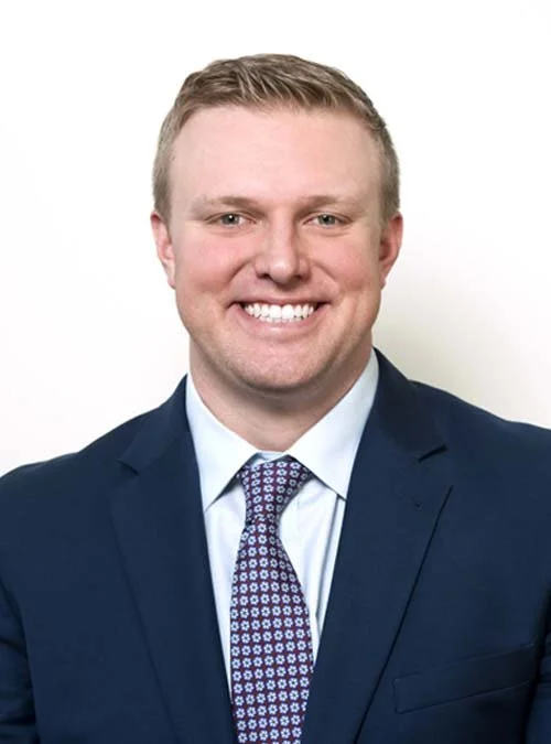 A young man in a dark suit, white shirt, and patterned tie, smiling at the camera against a plain white background.