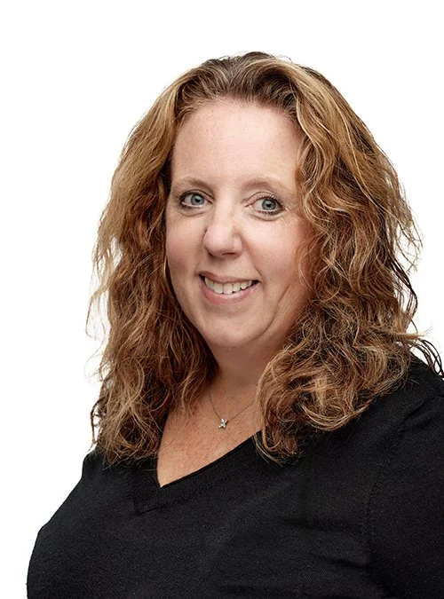 Portrait of a woman with curly red hair, smiling, wearing a black top and a necklace, against a white background.