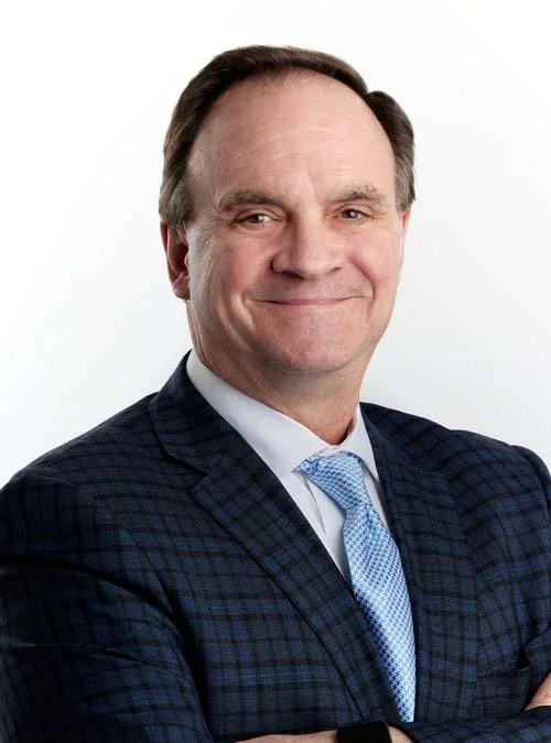 A middle-aged man with dark brown hair in a professional suit, smiling with arms crossed, against a white background.