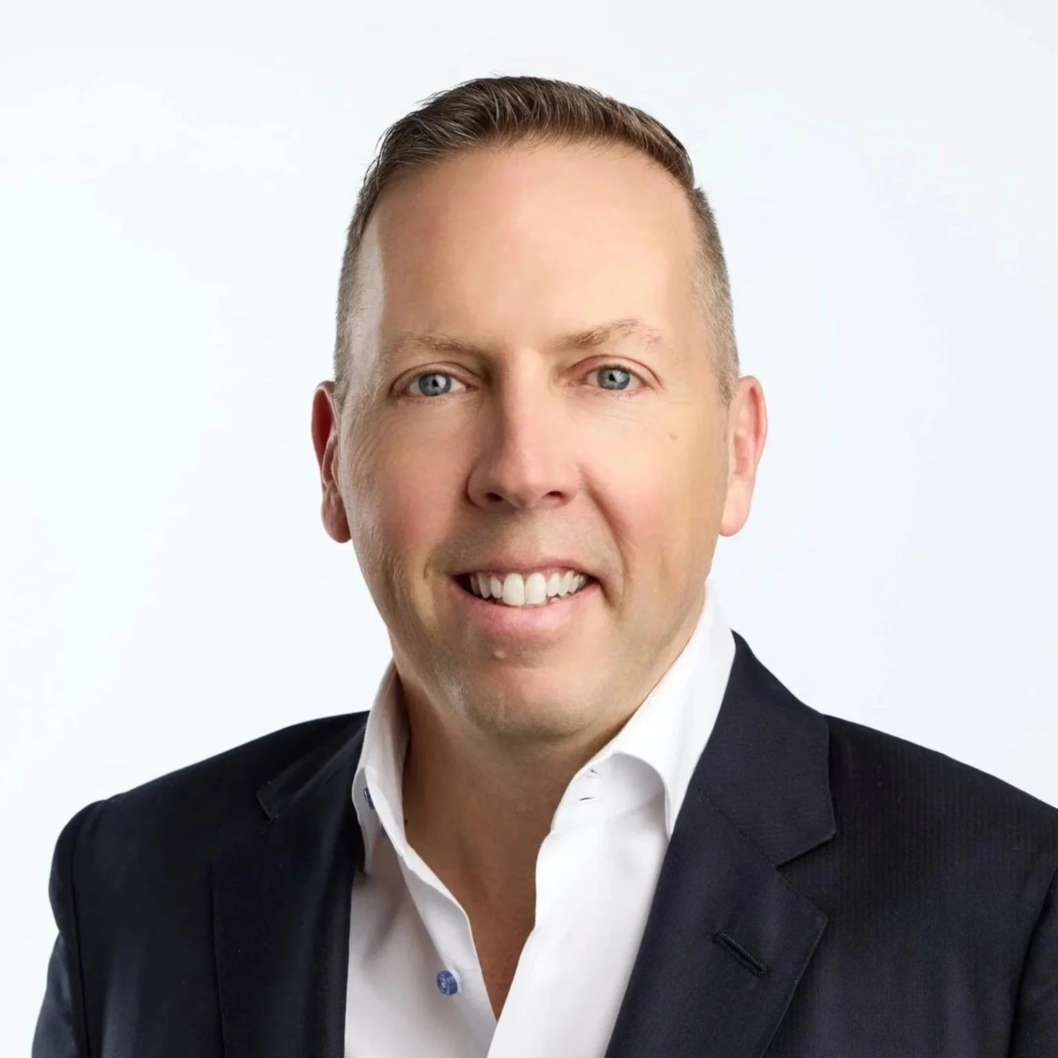 Professional headshot of a man with short light brown hair, blue eyes, wearing a white collared shirt and dark blazer, smiling against a plain white background.