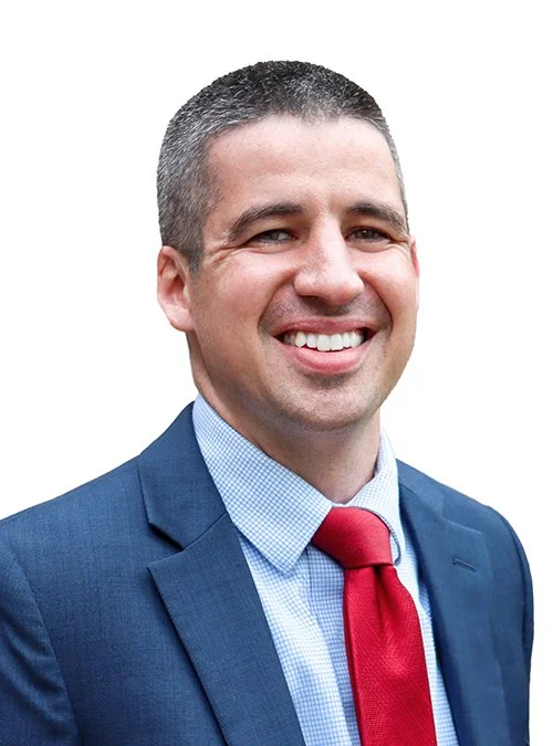 A smiling man in a blue suit with a light blue shirt and red tie, against a white background.