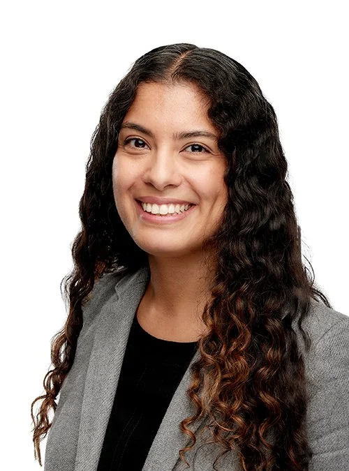 A professional woman with long, curly hair smiling at the camera, wearing a gray blazer and black top.