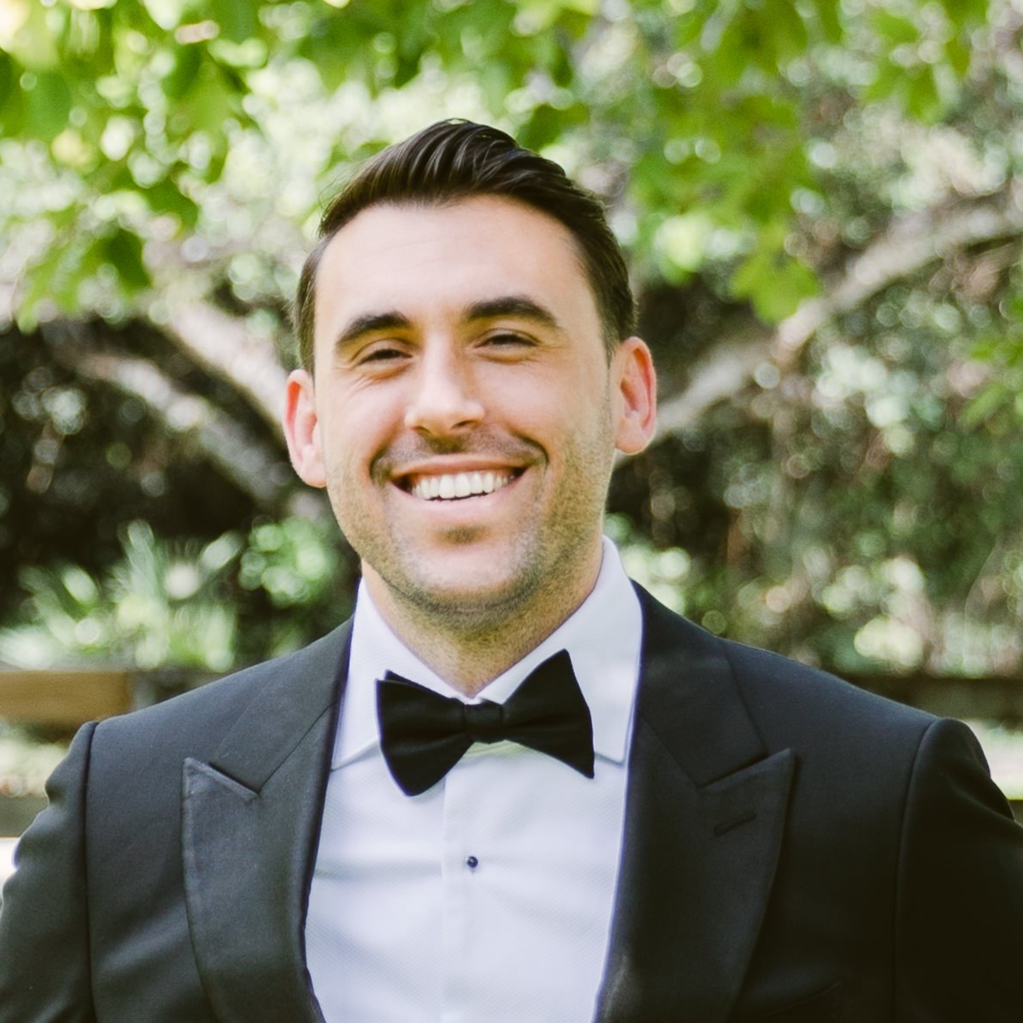 A smiling man in a tuxedo with a bowtie, standing outside with green foliage in the background.