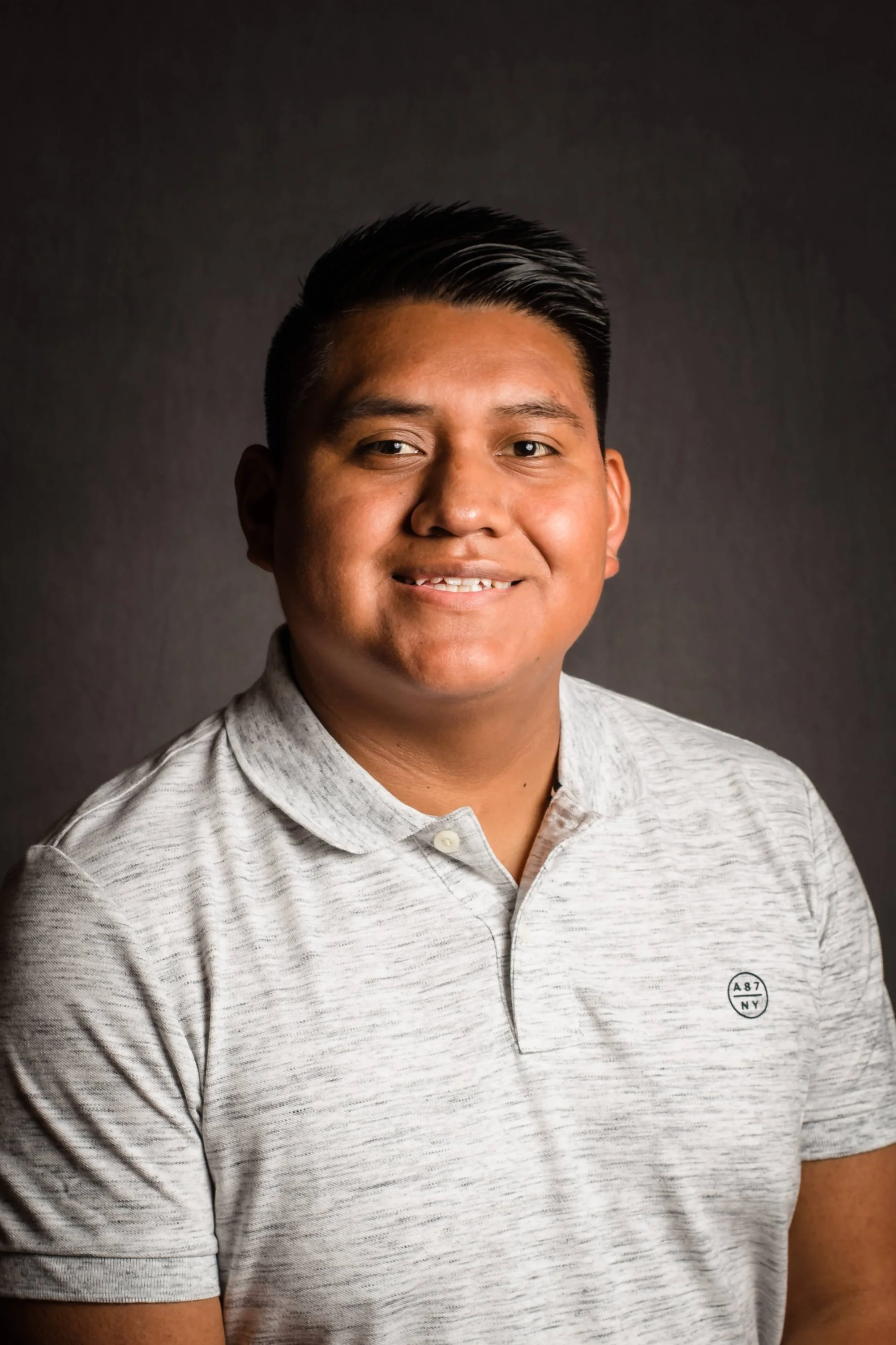 A young man with short dark hair smiling, wearing a light gray collared shirt, against a dark background.