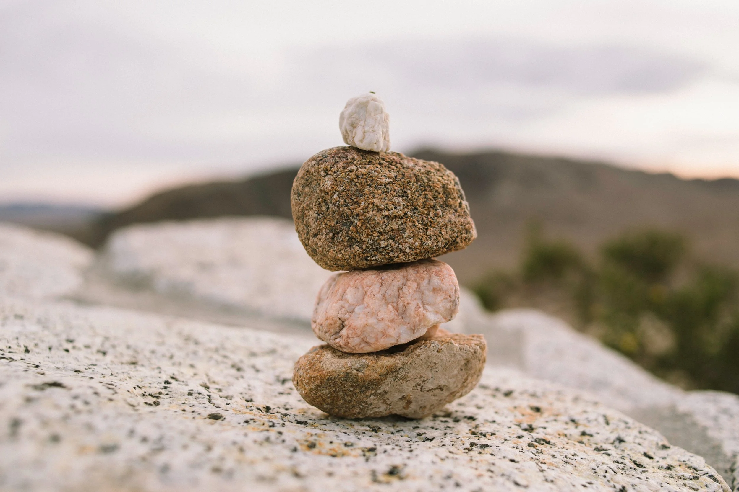 Stacked rocks on a sandy surface with a blurred landscape and cloudy sky in the background.