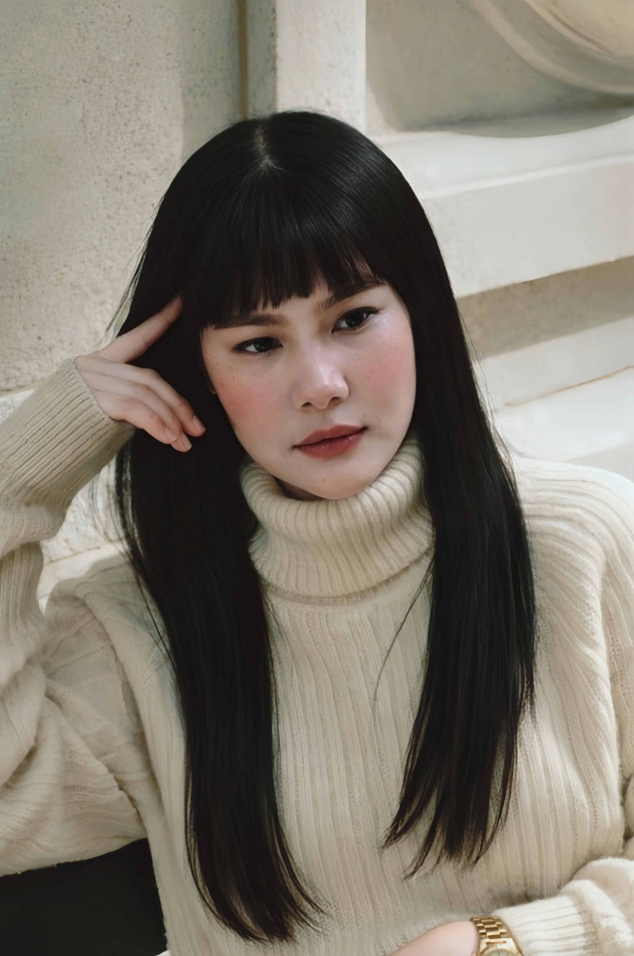 A woman in a cream-colored turtleneck sweater with long black hair and a gold watch, sitting against a beige wall, resting her head on her left hand.