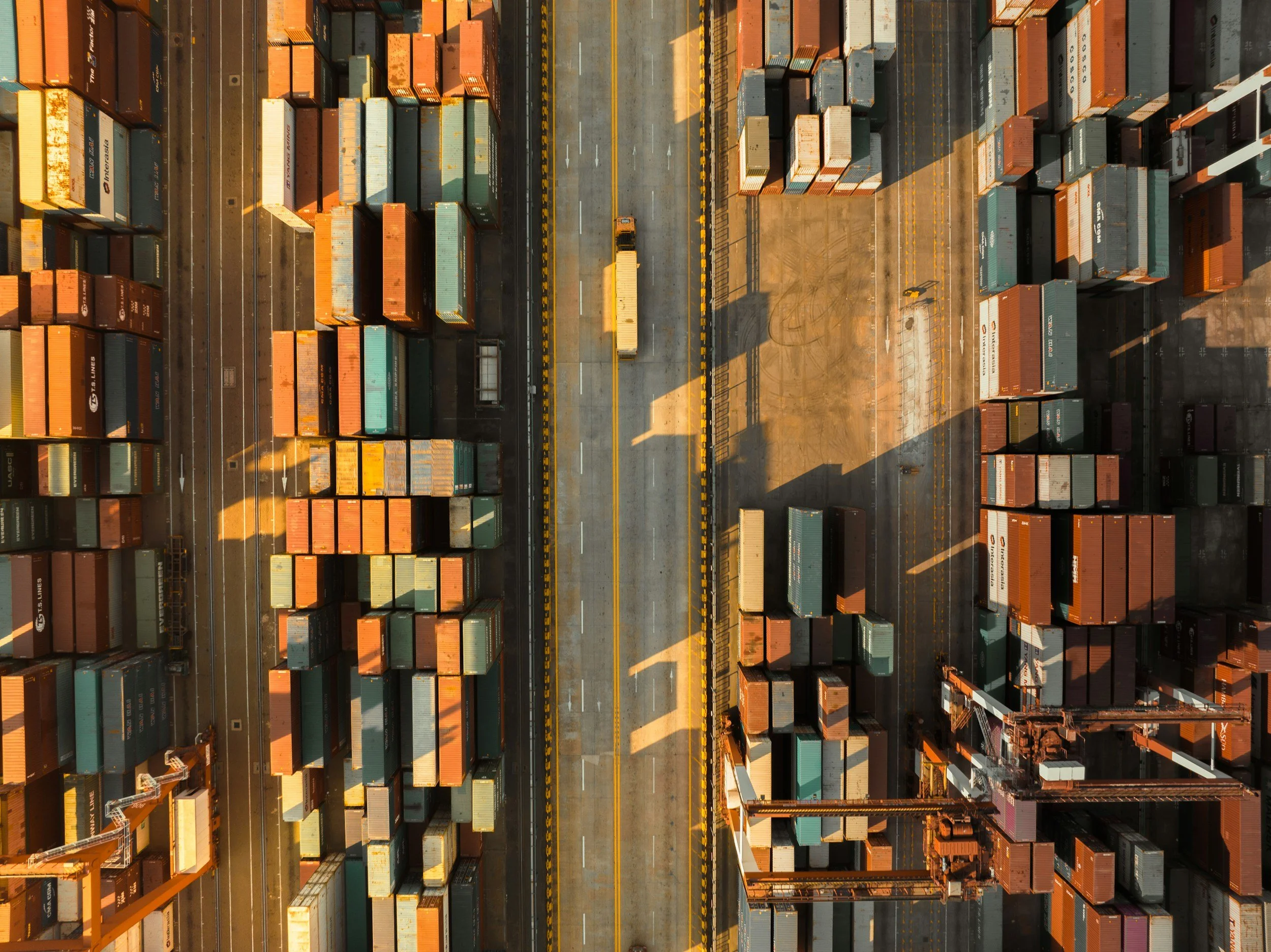 An aerial view of a shipping container yard with containers stacked on both sides and a small truck on a road in the middle, with shadows cast by nearby cranes and trees.
