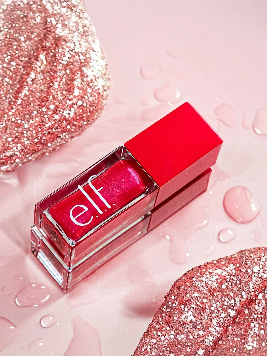 A bottle of elf liquid lipstick in a pink shade on a pink surface with water droplets, flanked by glittery pink objects.