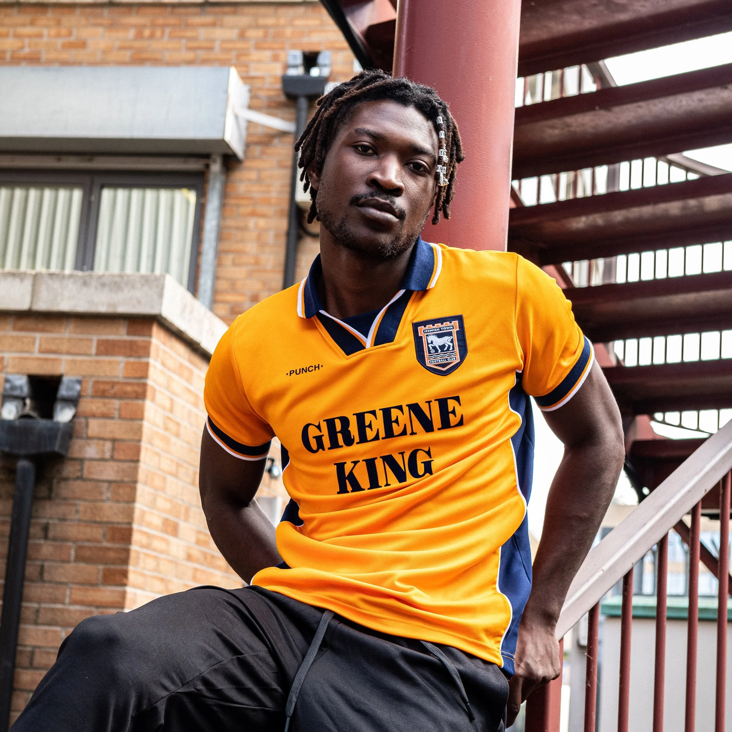 Young man in yellow sports jersey with 'GRENE KING' printed on front, leaning against a pole outside near brick buildings and a staircase.
