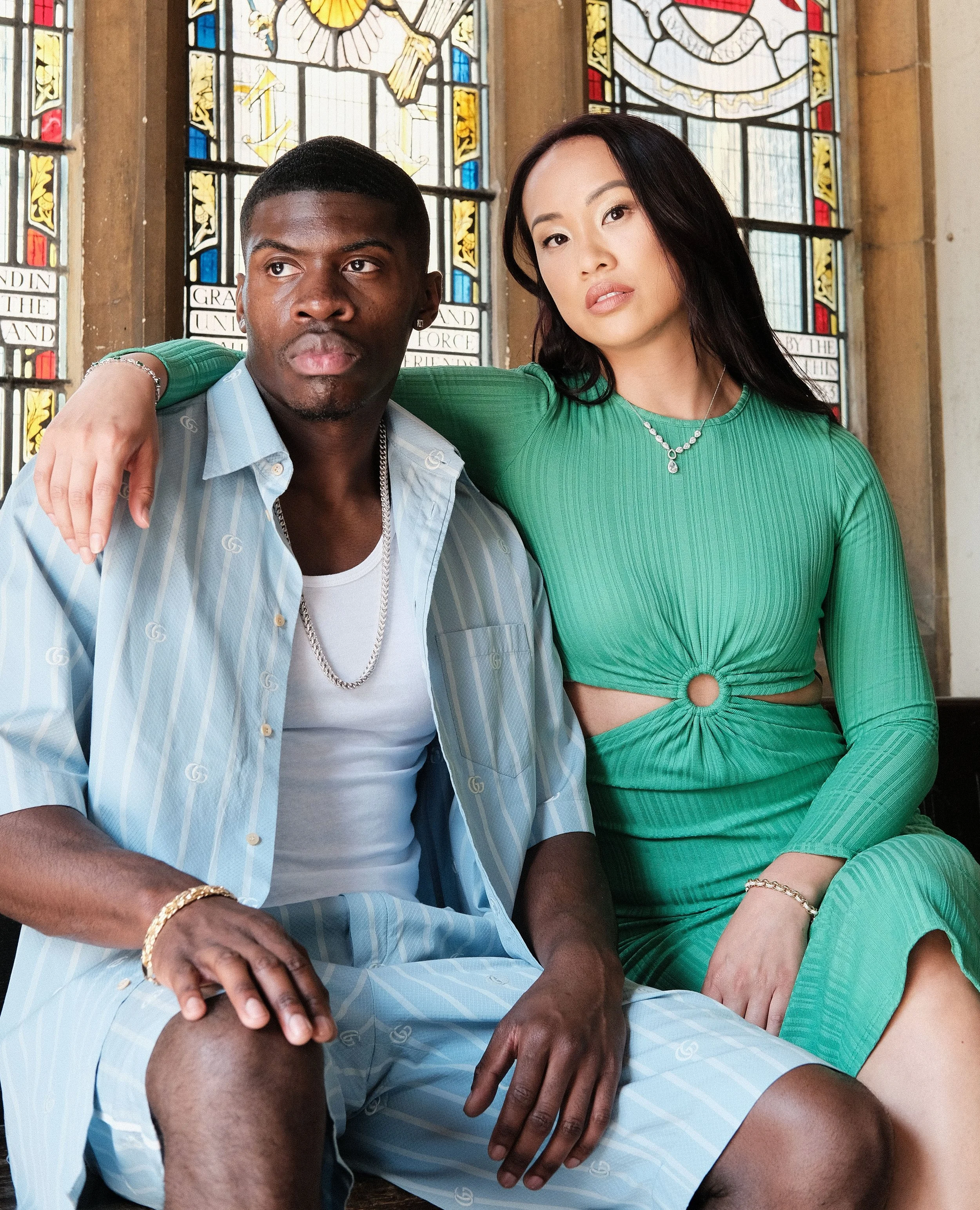 A man and a woman sit together inside a church with stained glass windows behind them. The man wears a light blue striped shirt and matching shorts, with jewelry including a chain and bracelet. The woman wears a green dress with cutouts, and she has a necklace and bracelet. The man has his arm around the woman’s shoulders.