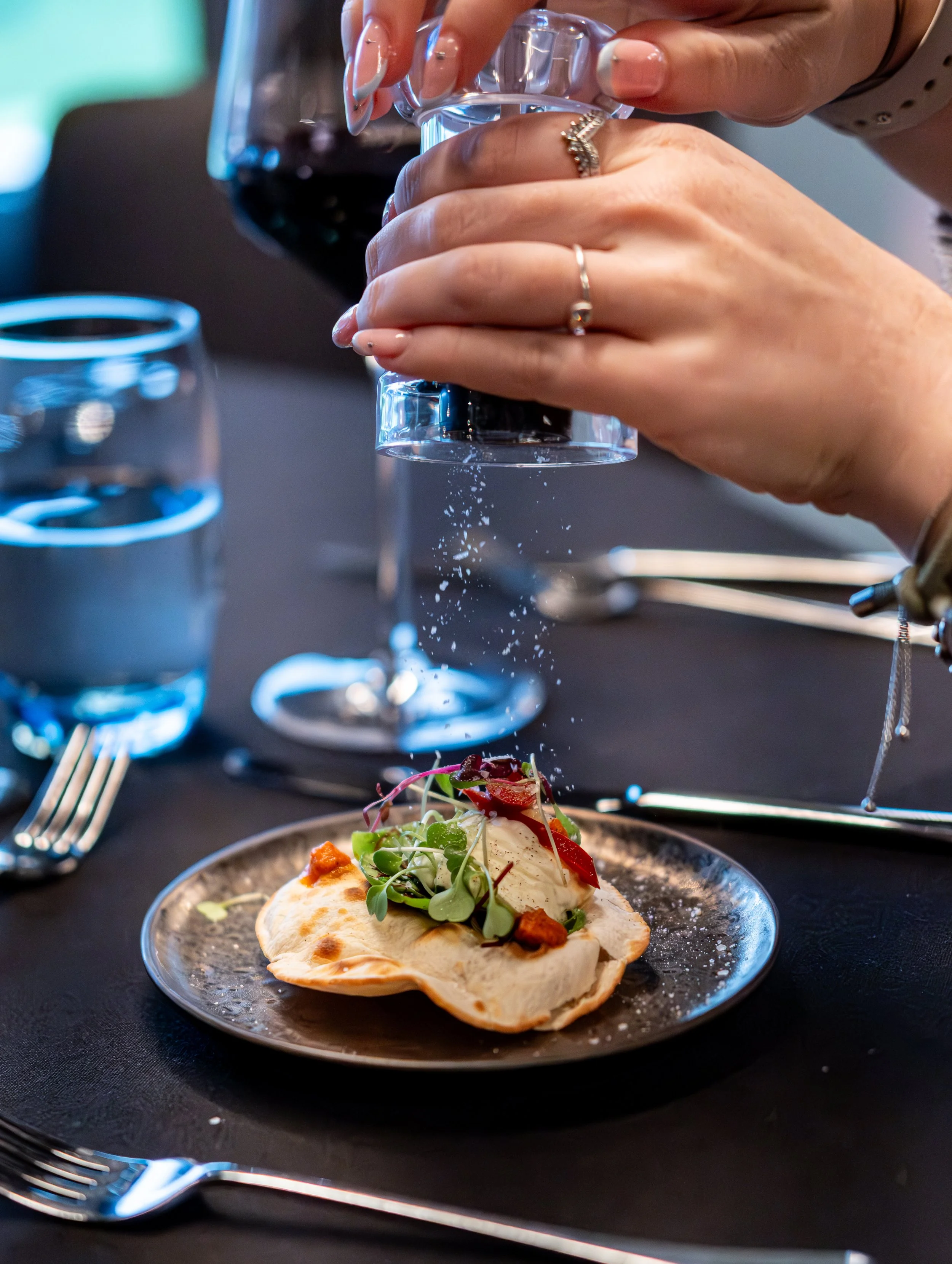 Person seasoning a plate of gourmet food with salt, with a glass of red wine and a glass of water on a black table.