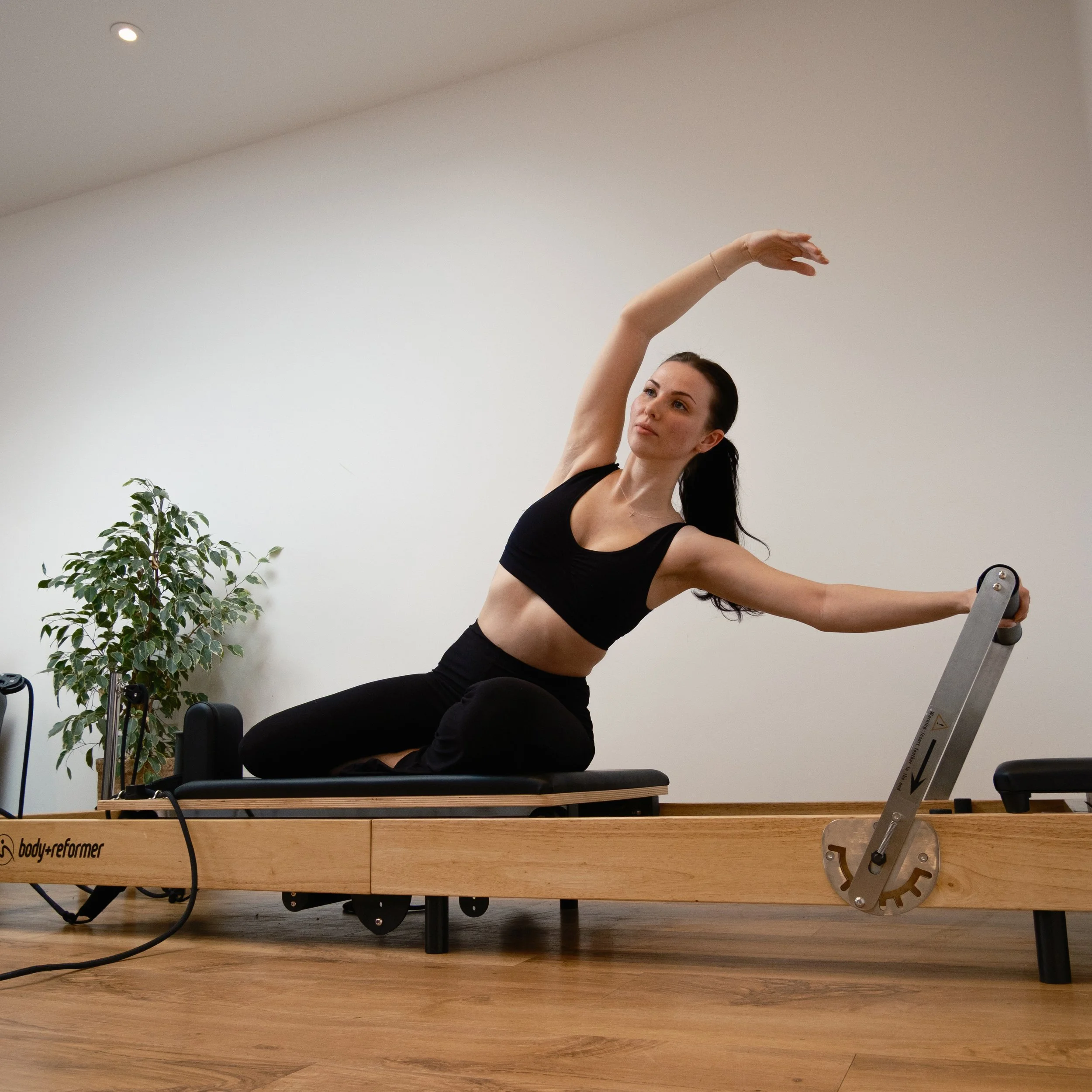 Woman exercising on a Pilates reformer machine in a studio, stretching with arms raised overhead and side.