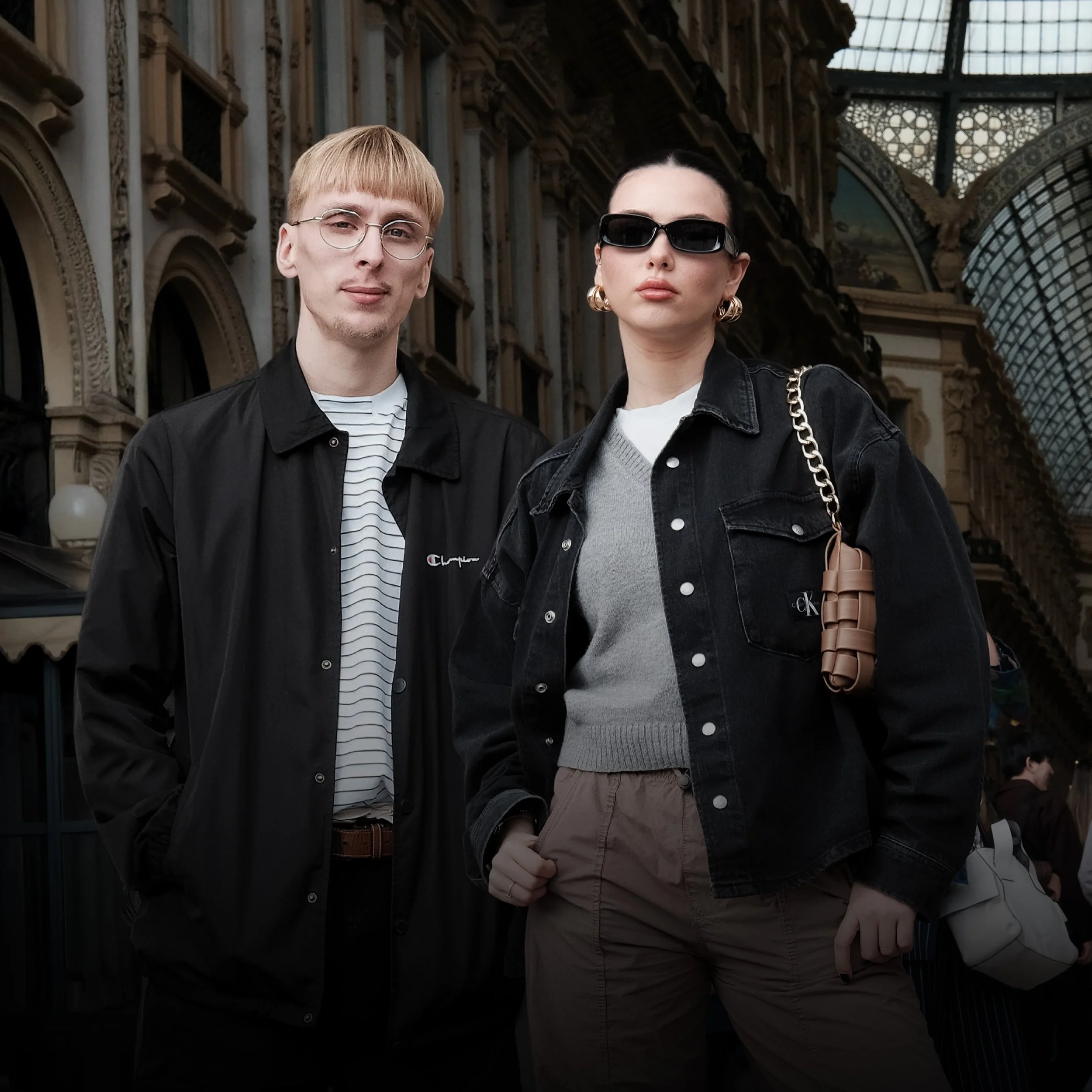 Two young adults, a man and a woman, standing indoors in front of ornate architecture with glass ceilings. The man wears glasses, a black jacket, and a striped shirt. The woman wears sunglasses, a black denim jacket, and a gray sweater, carrying a beige woven handbag.