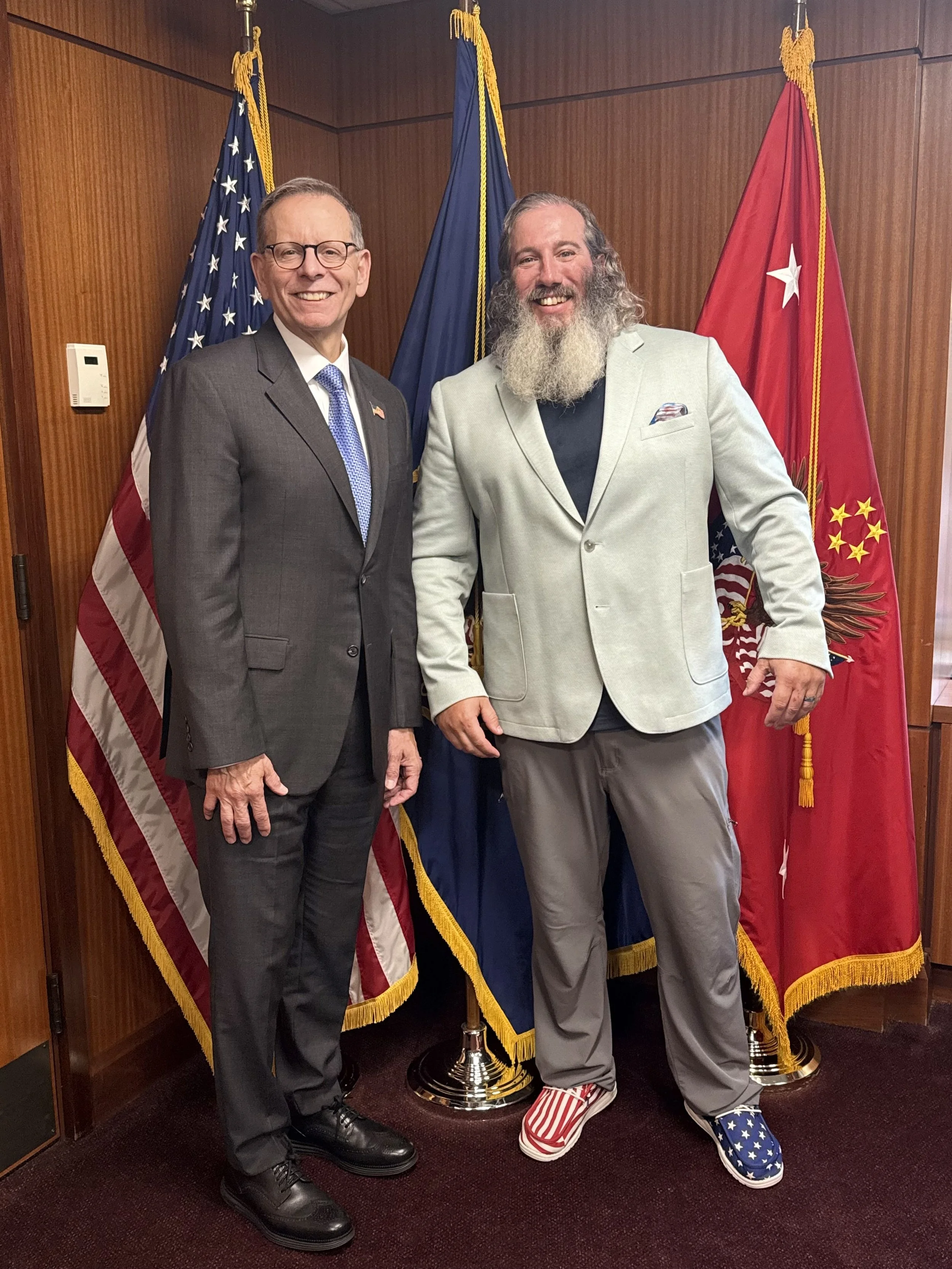 Two men standing in front of American and military flags, smiling, in a wood-paneled room. The man on the left wears a gray suit, glasses, and a blue tie. The man on the right has a long beard, gray hair, gray suit, and distinctive American flag-them