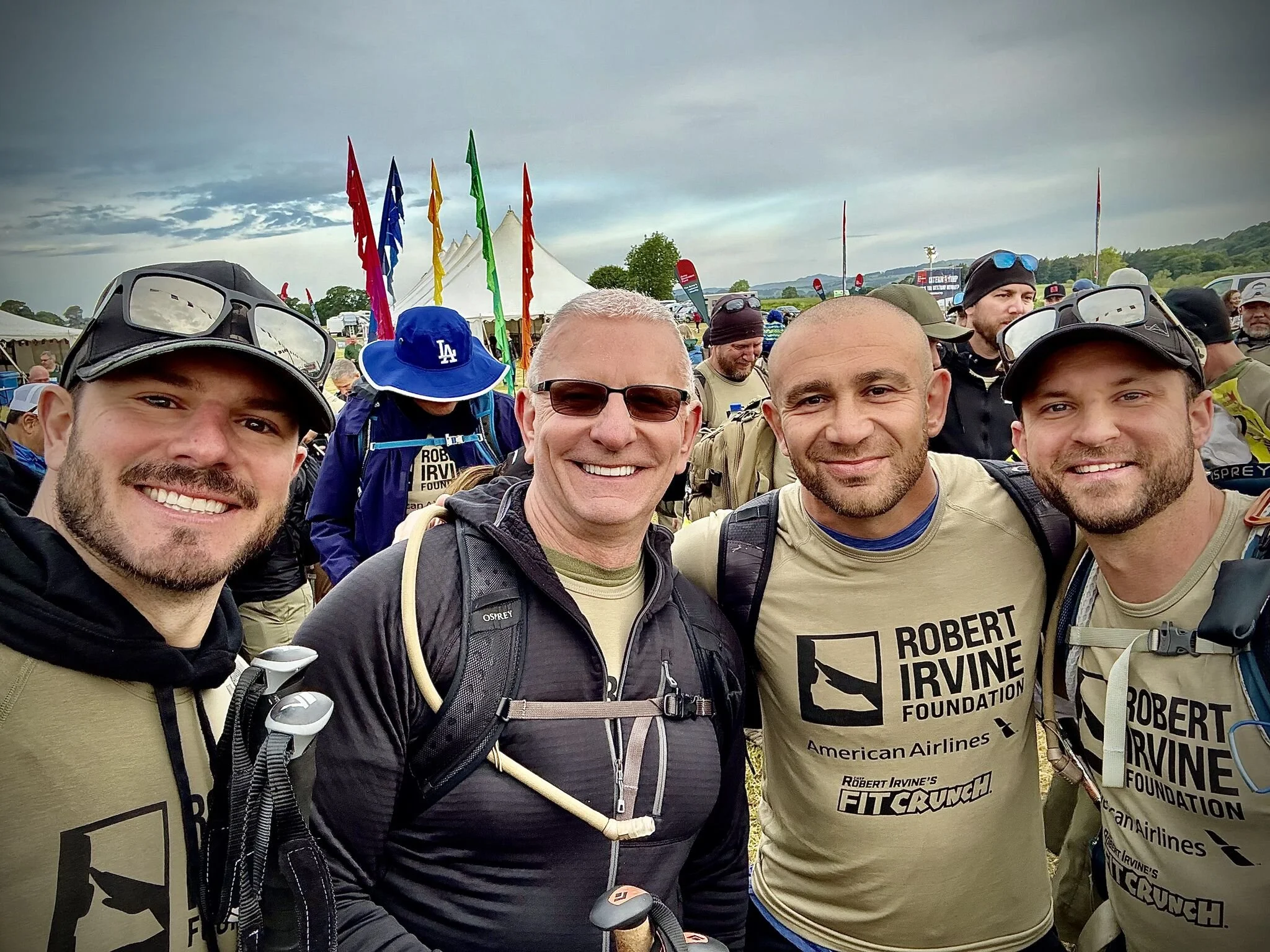 Group of smiling men outdoors at a charity or awareness event, with colorful flags and tents in the background, wearing athletic clothing and backpacks.