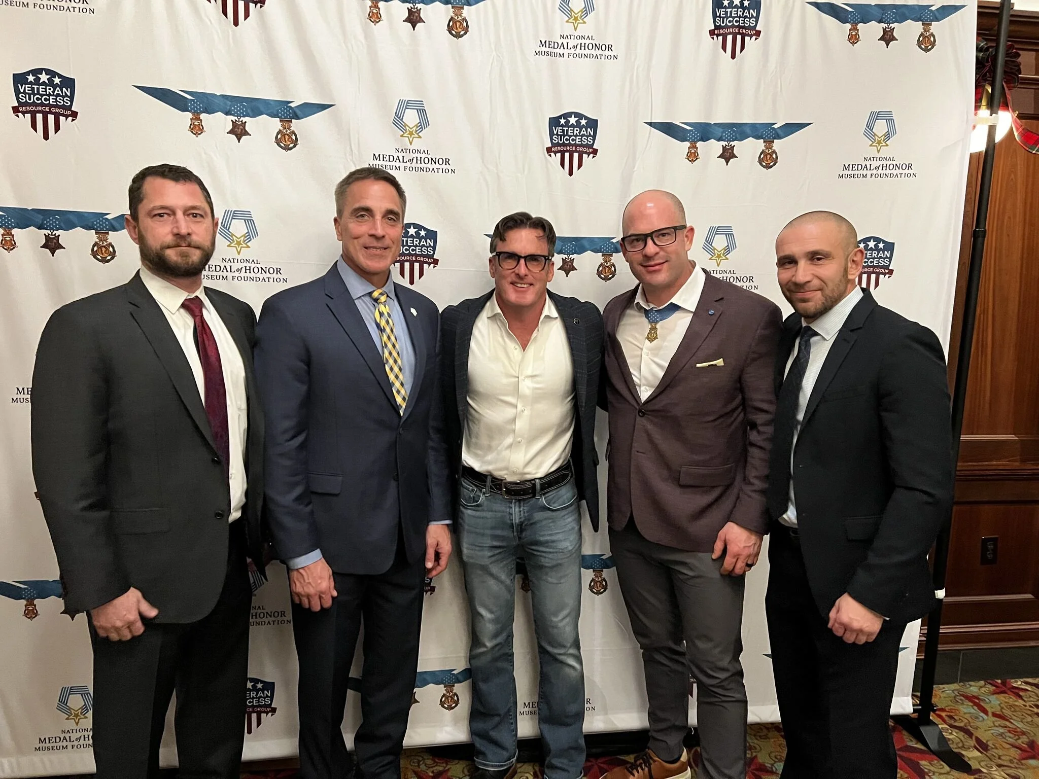 Five men in formal suits standing in front of a backdrop with logos for the National Medal of Honor Museum Foundation and Veteran Success Resource Group, smiling at the camera.