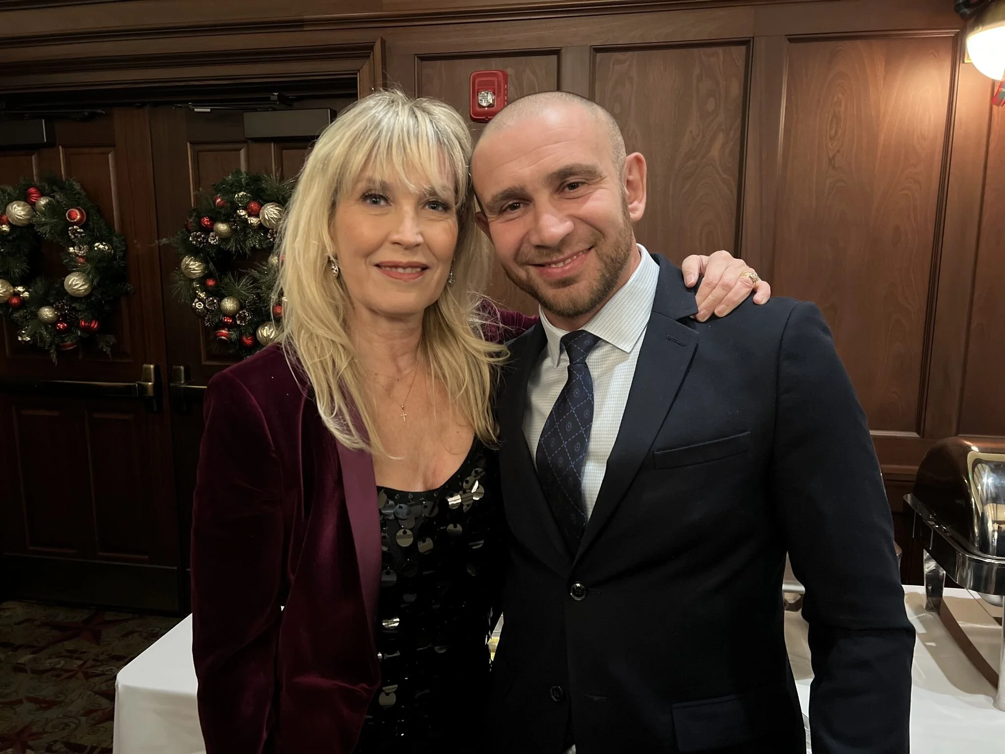 A woman with blonde hair and a man with a shaved head and beard smiling and posing together in formal attire in a room with wooden paneling and Christmas wreaths decorated with gold and red ornaments in the background.