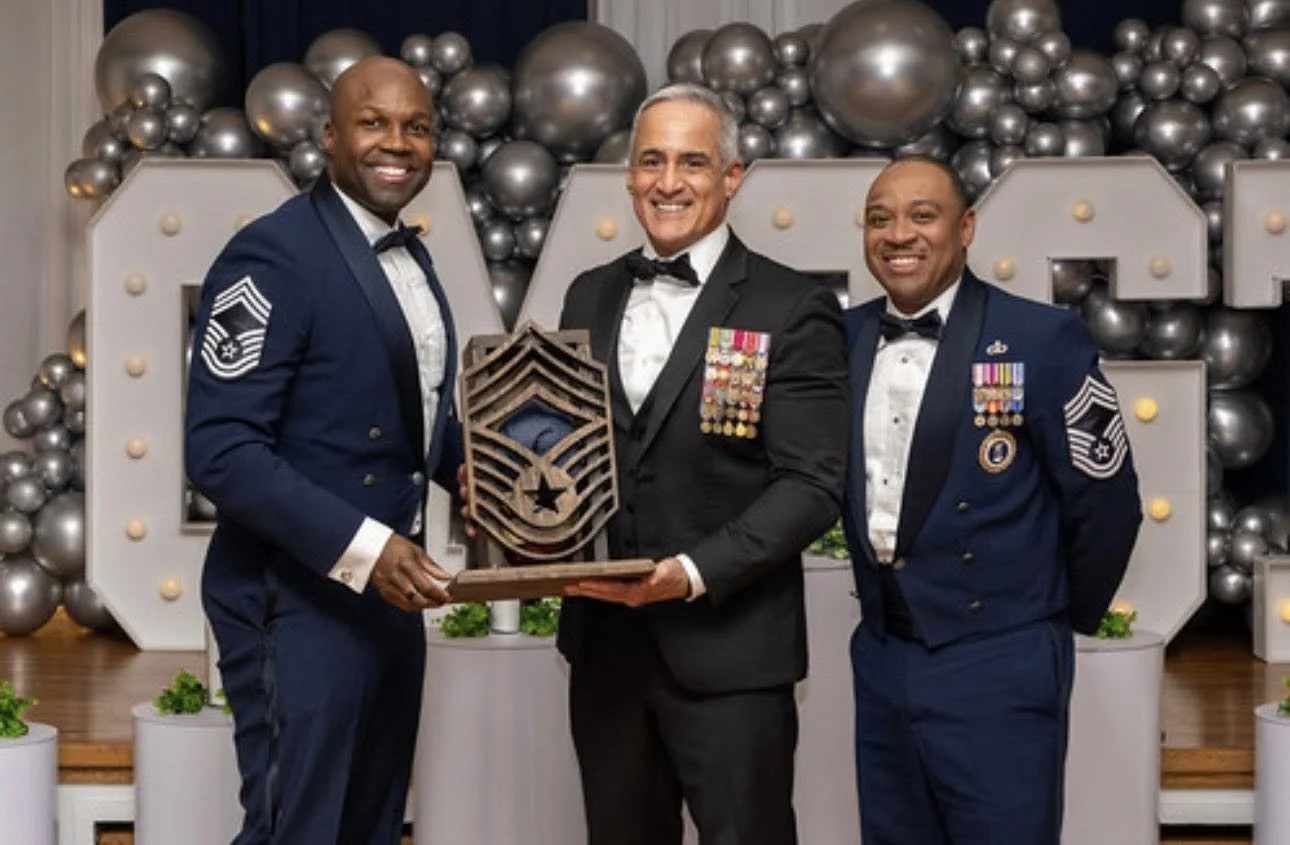 Three men in military dress uniforms and the man in the middle in a black tuxedo holding a framed military rank insignia at a decorated event with silver balloons and large illuminated letters.