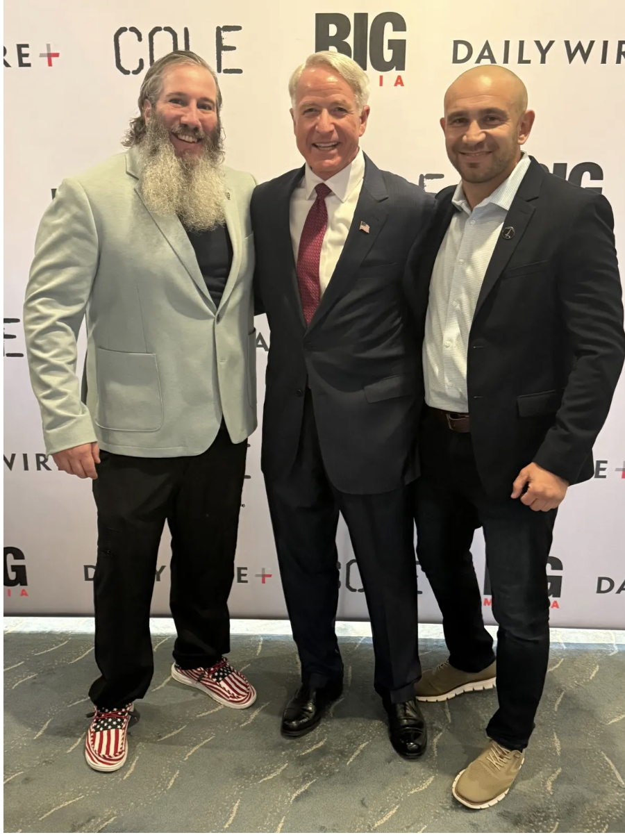 Three men in suits posing for a photo at an event with a backdrop displaying logos such as 'CONE,' 'BIG,' and 'DAILY WIRE.'