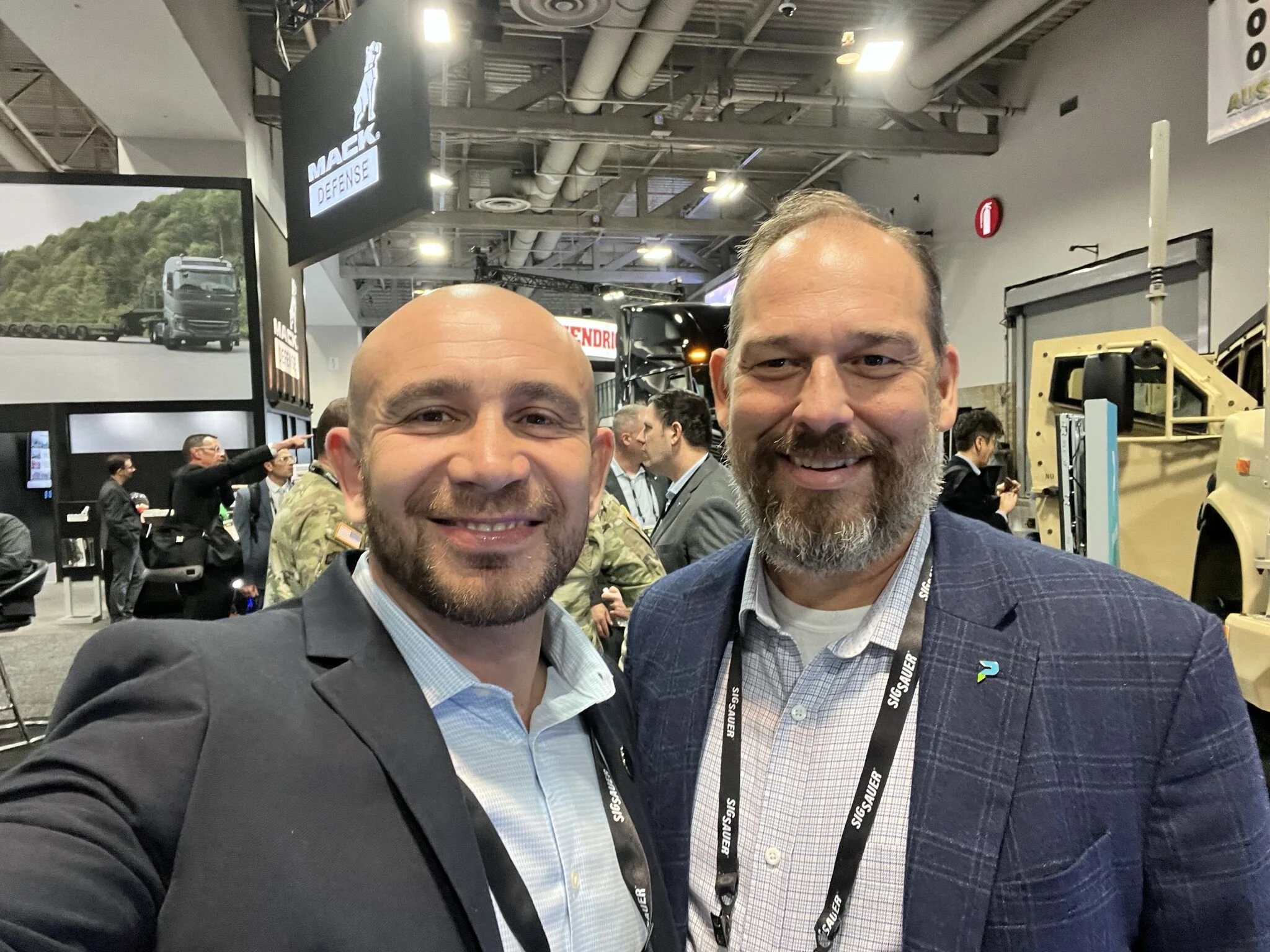 Two smiling men taking a selfie at a trade show or conference, standing in front of various exhibition booths and displays, with people chatting and looking at products and presentations in the background.