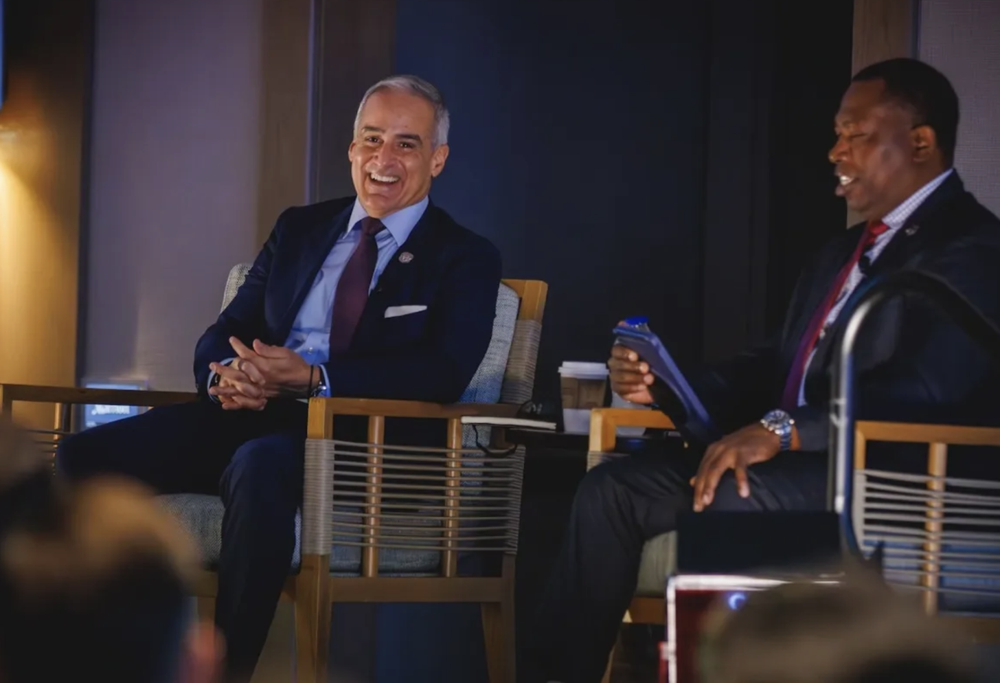 Two men in suits sitting on stage, smiling and engaging in conversation during an event or panel discussion.
