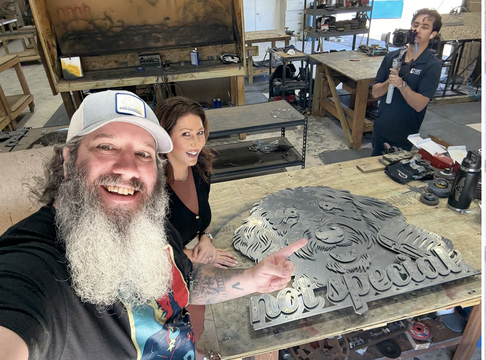 Three people in a workshop, with a large metal lion and text on a wooden table in front of them. One man with long gray beard and cap, smiling and pointing at the metal art piece, a woman beside him smiling, and a man in the background filming with a