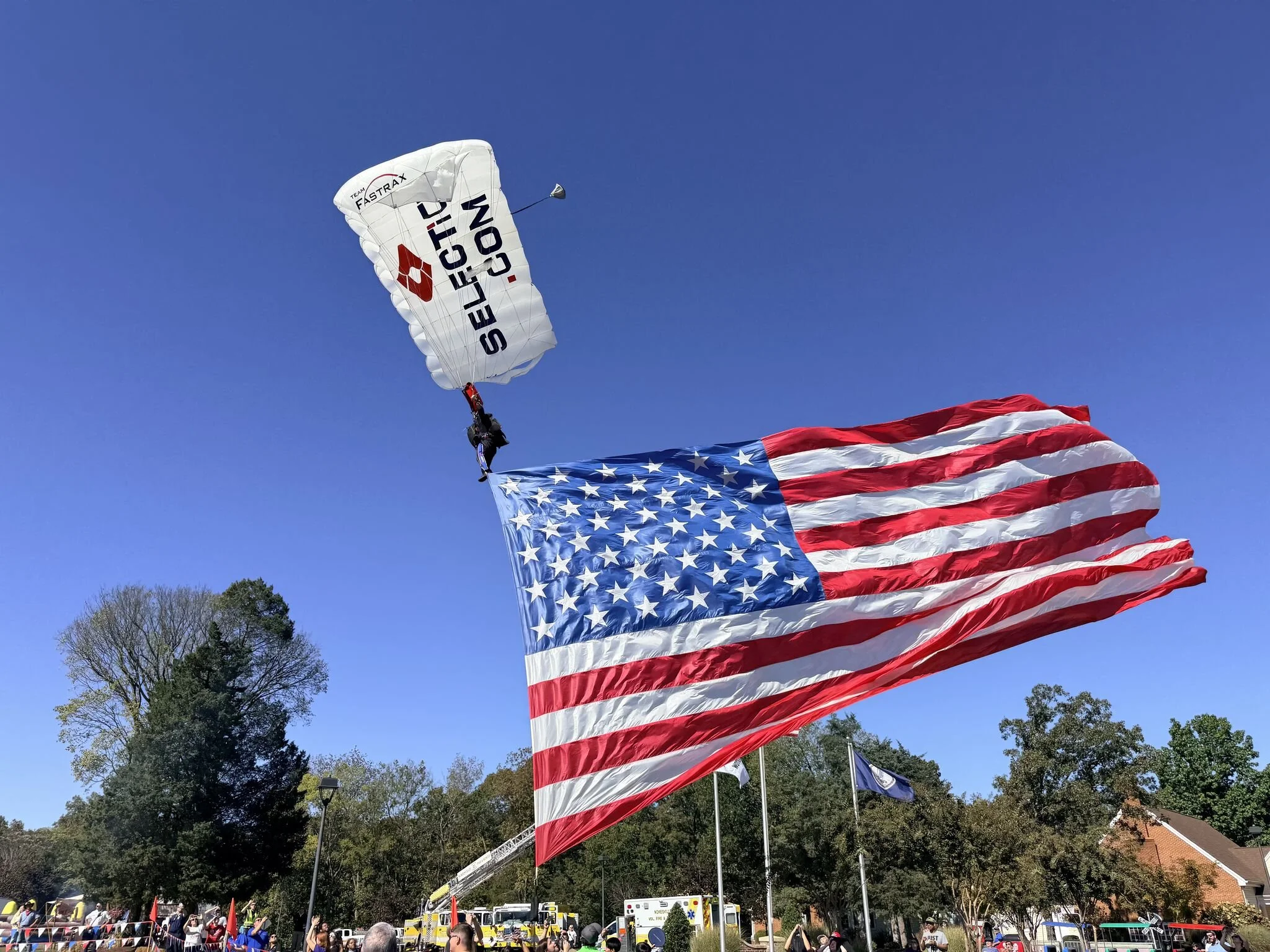 A person skydiving with a parachute that has the logo of Equipo EIC and the words 'SAVE IT IS COM' printed on it, holding an American flag during a public event.