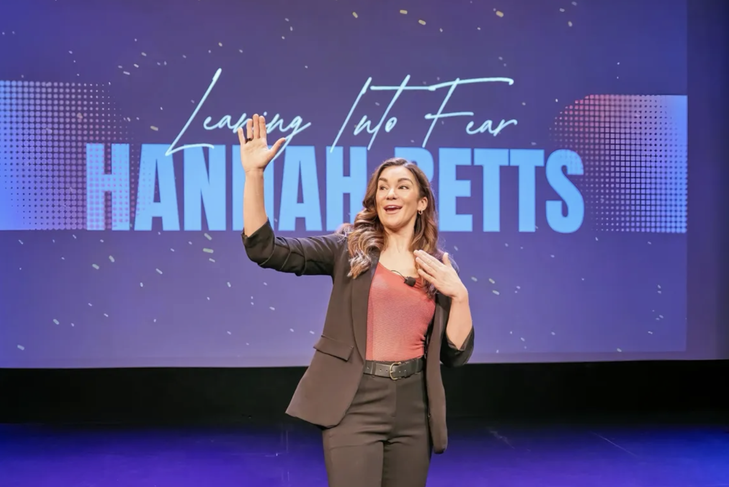 A woman standing on stage, gesturing with her right hand and touching her chest with her left hand, in front of a screen that reads 'Learning Into Fear HANNAHETS'.