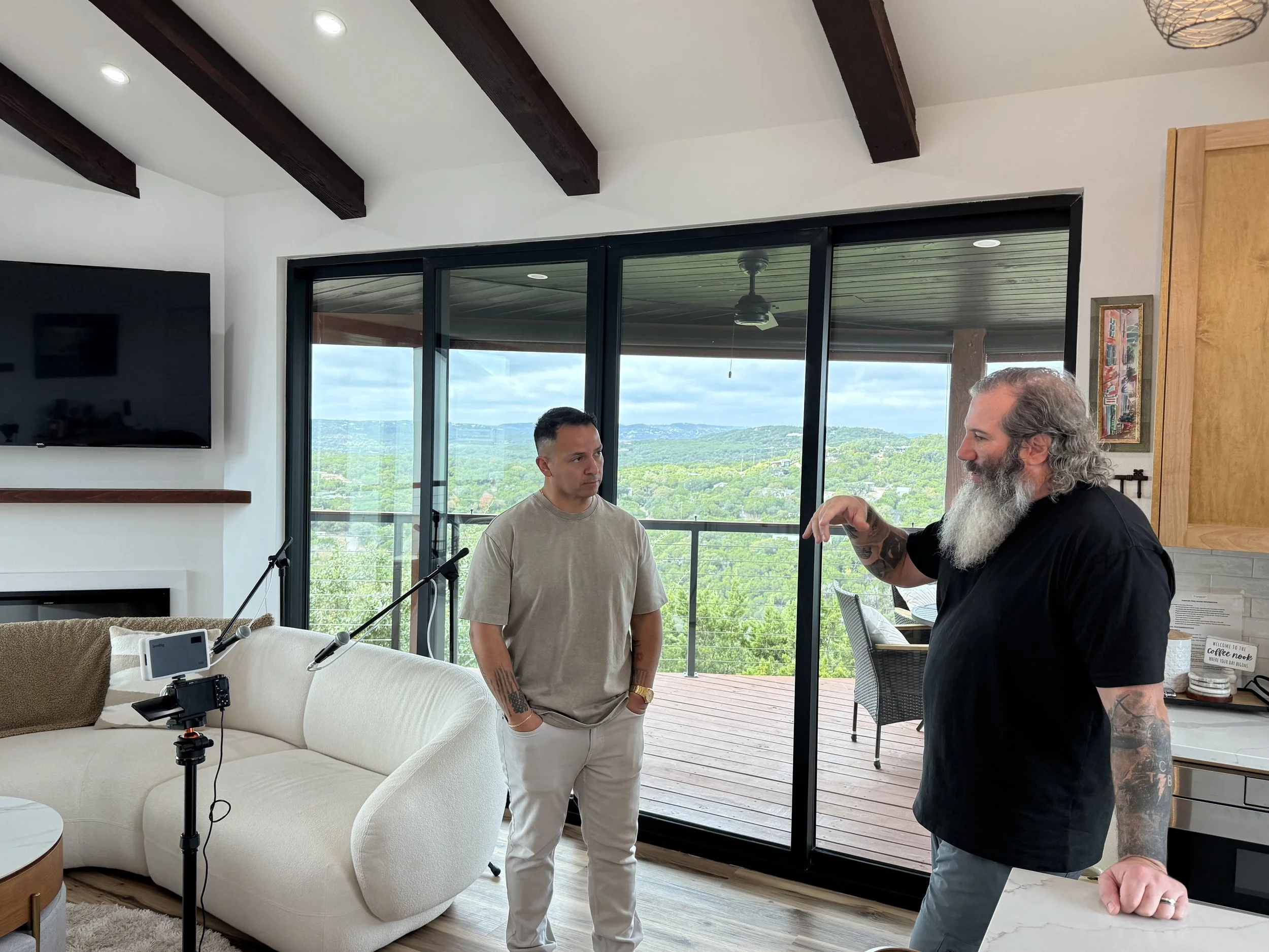 Two men are having a conversation inside a bright living room with large windows showing a scenic view of green hills and sky. One man has long gray hair and a beard, wearing a black t-shirt and tattooed arms. The other man has short dark hair, tatto