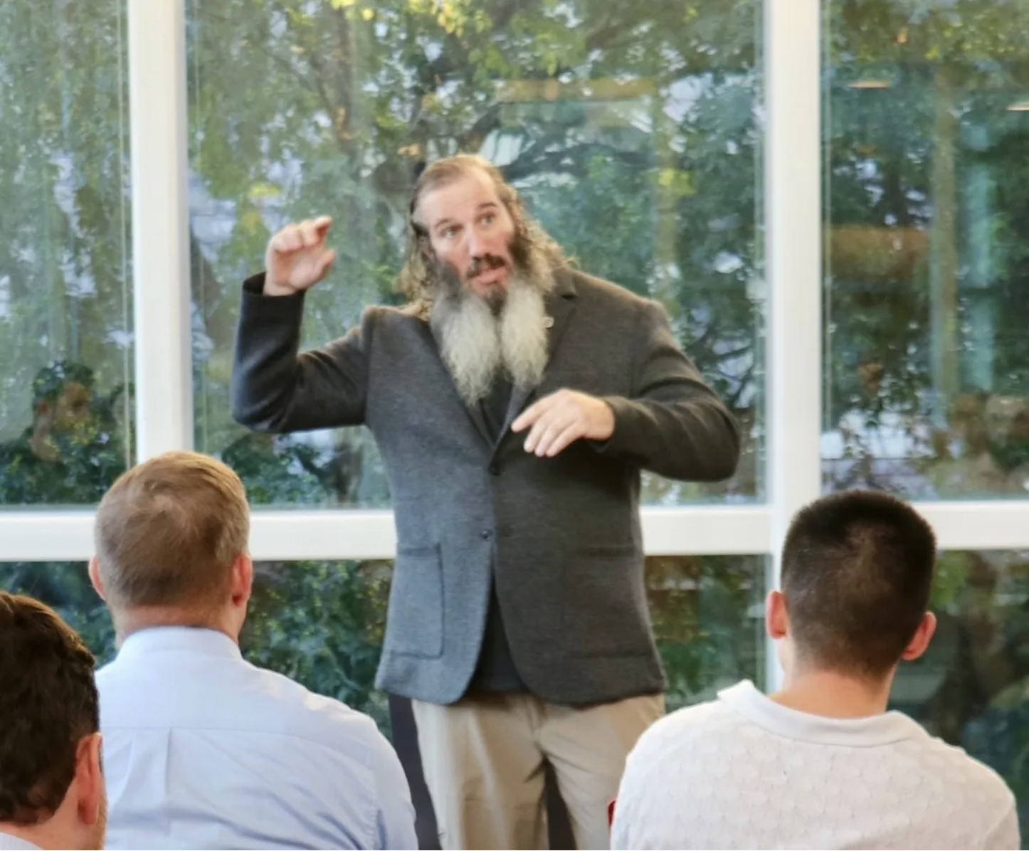 A man with long hair and a beard, wearing a dark jacket, is speaking or presenting in front of a group of people sitting indoors near large windows with trees outside.