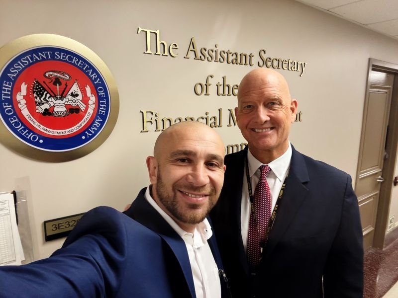 Two men in business attire smiling for a selfie in front of a sign that reads "The Assistant Secretary of the Financial Management" with an official seal of the Office of the Assistant Secretary of the Army Financial Management and Comptroller.