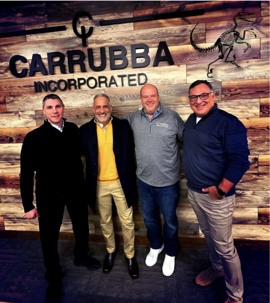 Four men standing in front of a wooden wall with a 'Carrubba Incorporated' sign and a scorpion graphic, smiling.