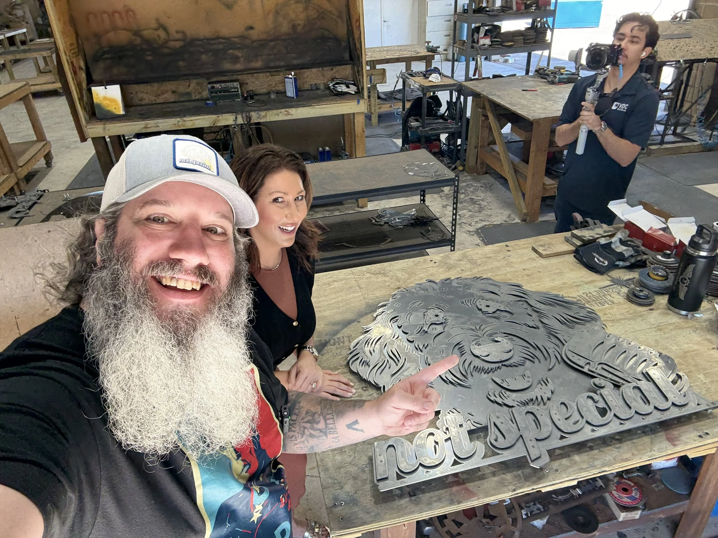 A group of three people in a workshop, two women and one man. The man in the foreground with a beard and hat is smiling and pointing at a metallic artwork of a lion's face on a workbench. The woman with brown hair, next to him, is smiling. The third 