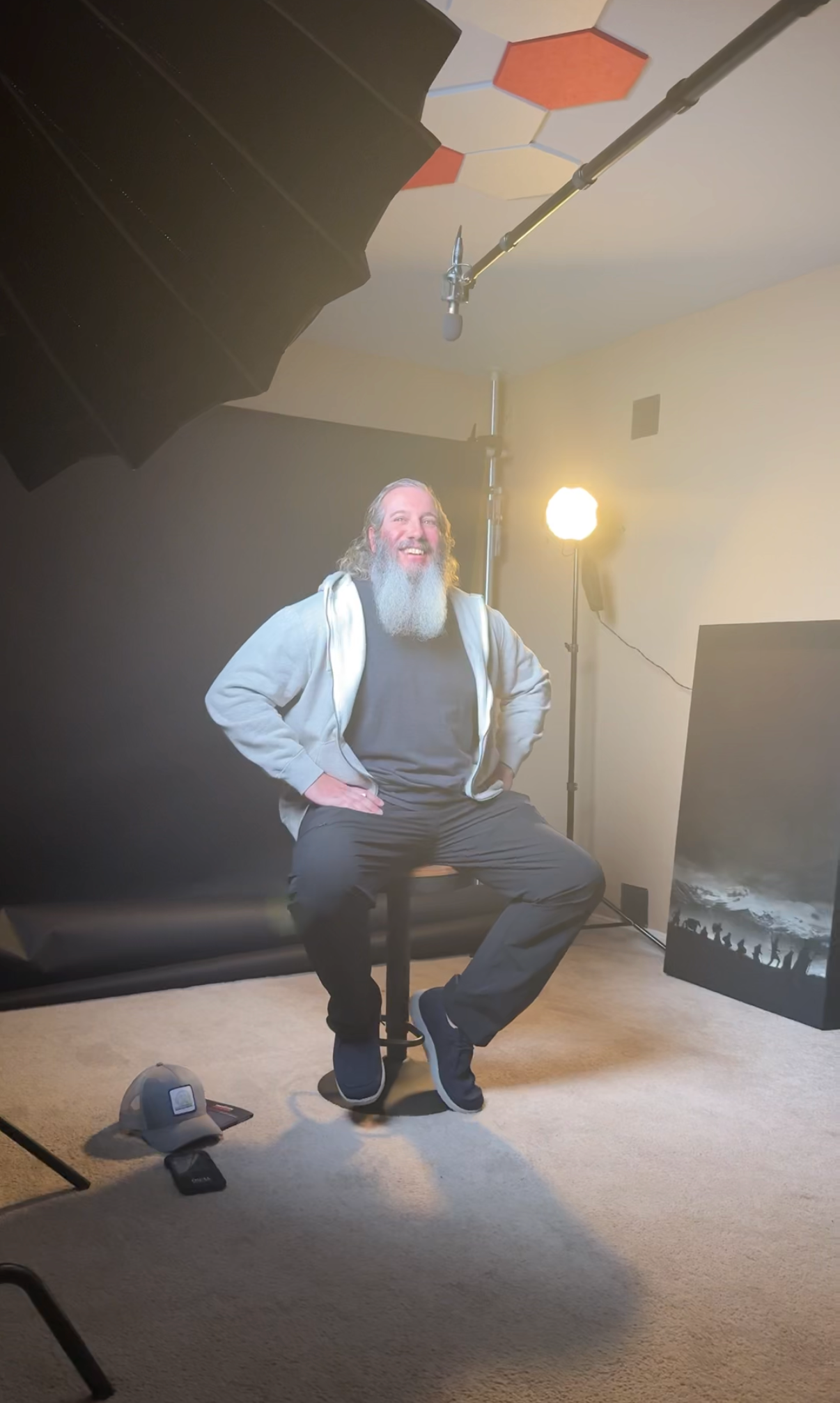 A man with long hair and a beard sitting on a stool in a photography or video studio. He is smiling and wearing a hoodie and dark pants. Behind him is a yellow studio light, and a large black backdrop hangs on the left. A camera is on a tripod on the