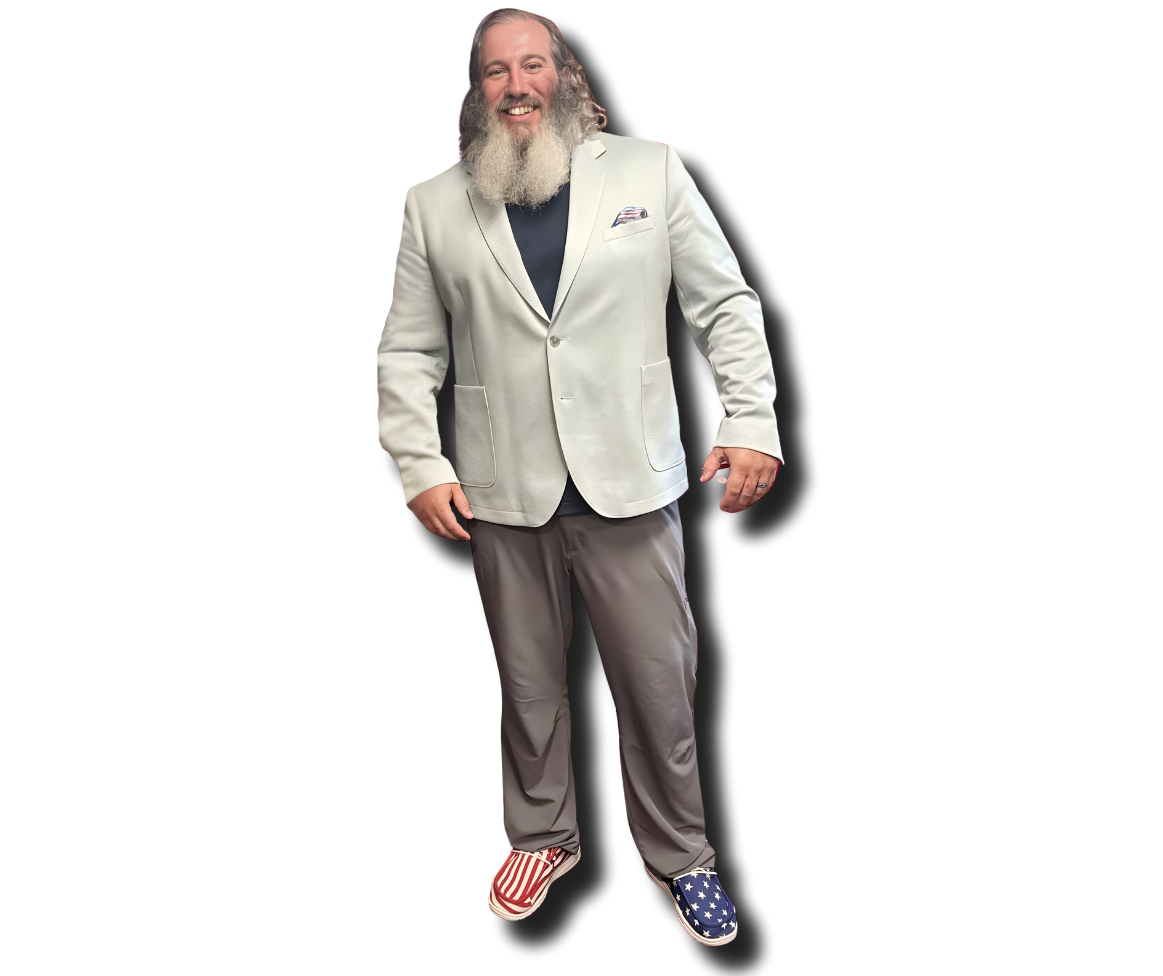 A man with a beard smiling, wearing a white blazer, dark shirt, gray pants, and American flag themed slippers.