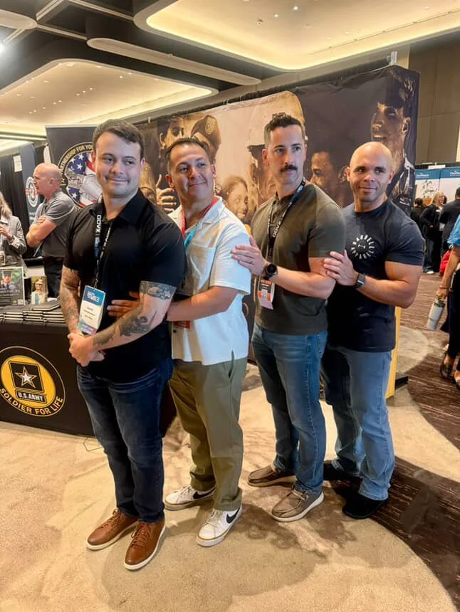 Four men standing in line, posing for a photo at an indoor event. They are in front of a booth with a U.S. Army Soldier for Life logo, and there are other booths and attendees in the background.