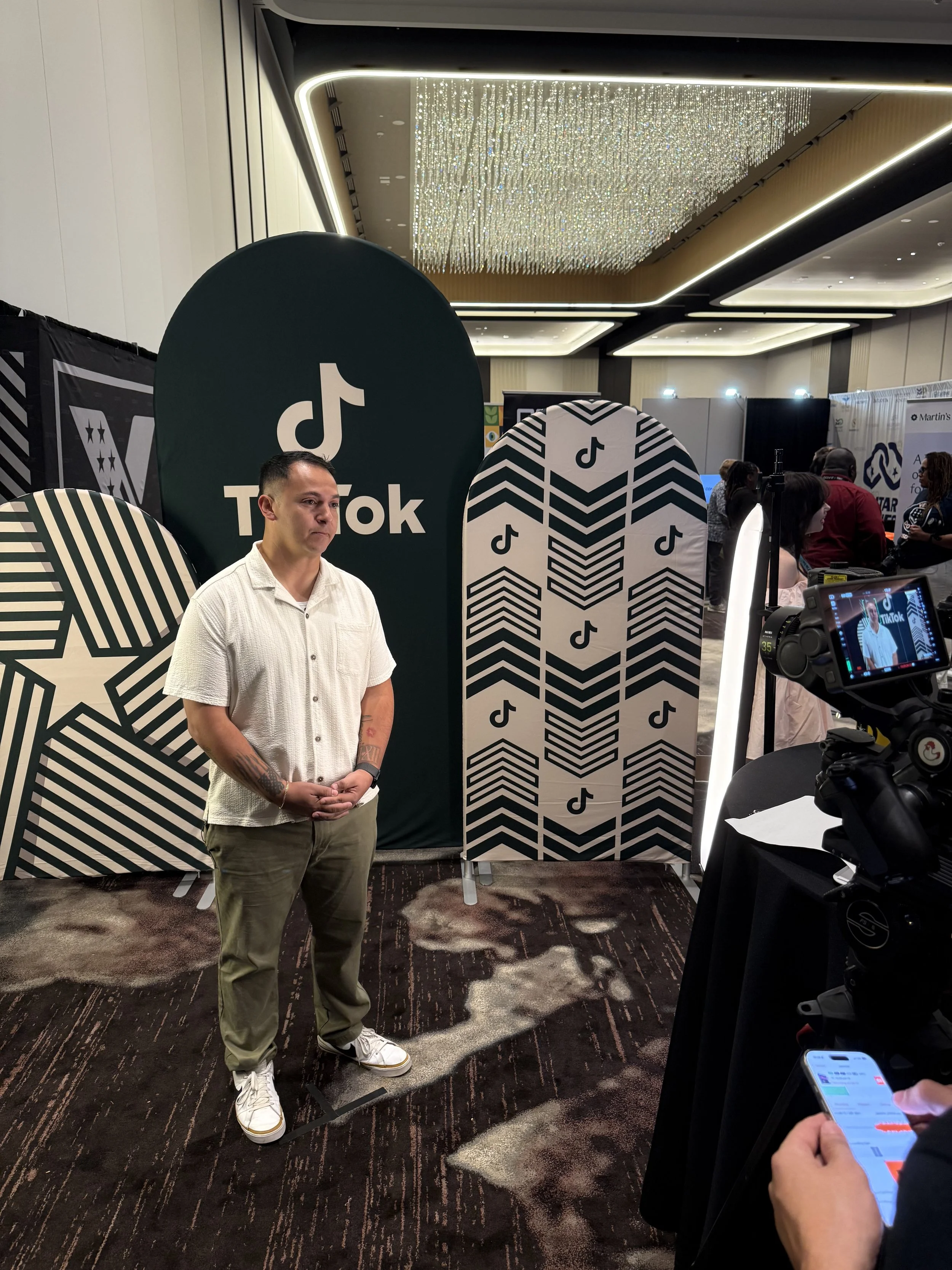 A man stands in front of a TikTok display at a conference or event. The display includes large cutouts with the TikTok logo and patterned designs. The environment has a modern, well-lit interior with a dramatic chandelier hanging from the ceiling. There are other attendees and booths visible in the background, and a person is recording with a camera while another is using a phone.