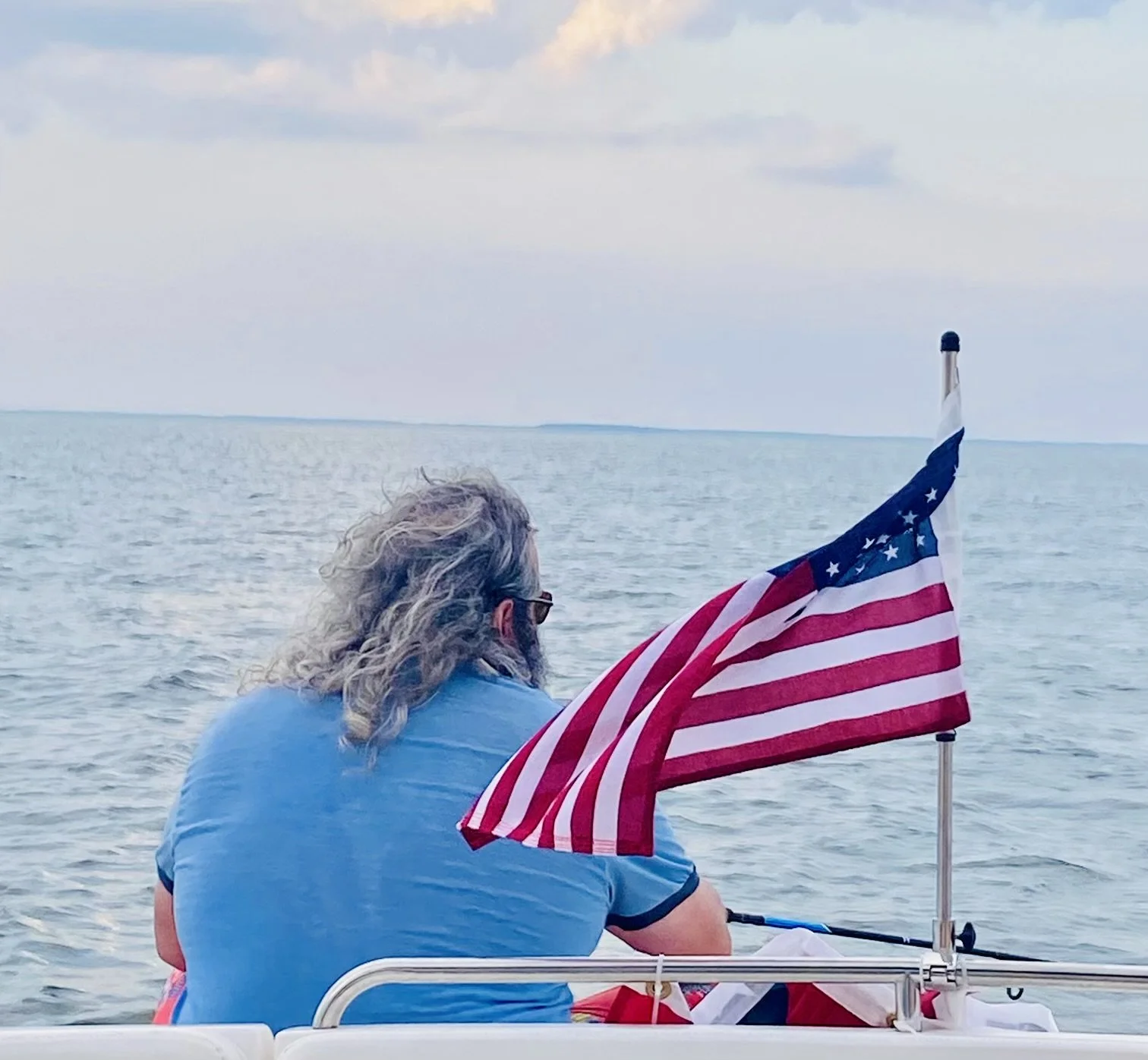A person with long gray hair and glasses sitting on a boat, looking out at the water with an American flag attached to the boat.