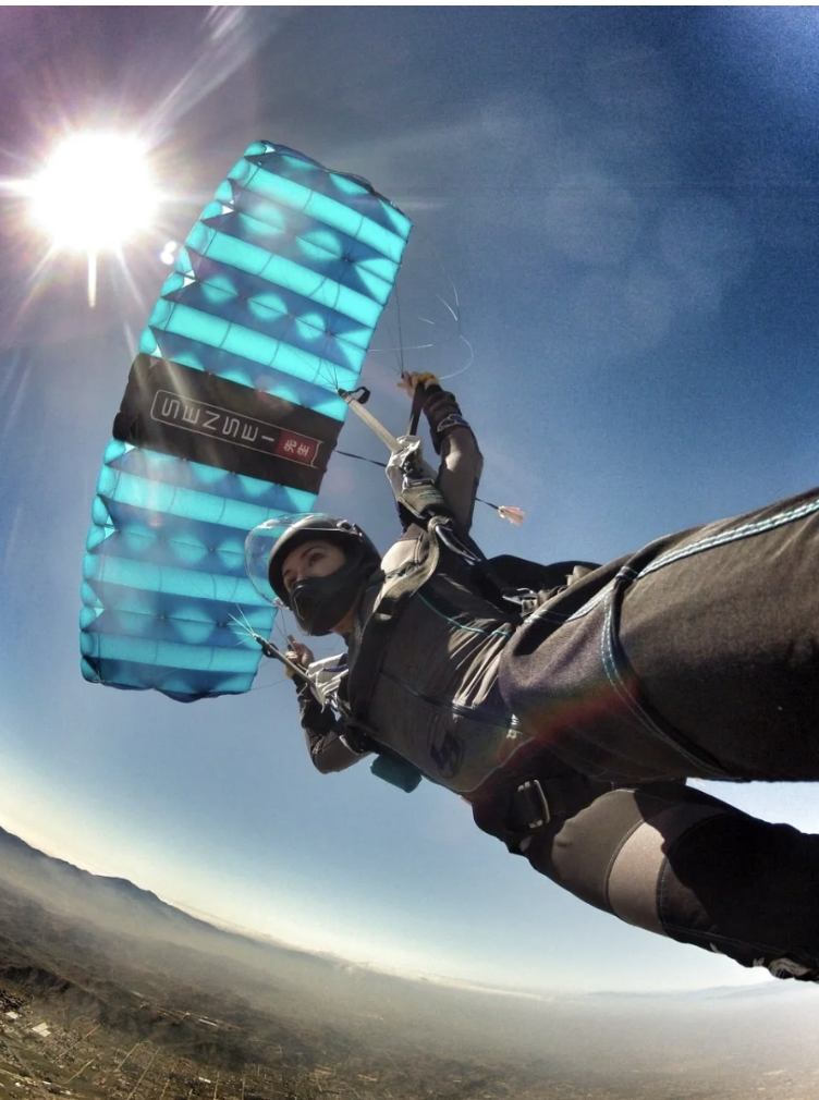 Person paragliding in the sky, with a blue paraglider, wearing a helmet and harness, and the sun shining brightly.