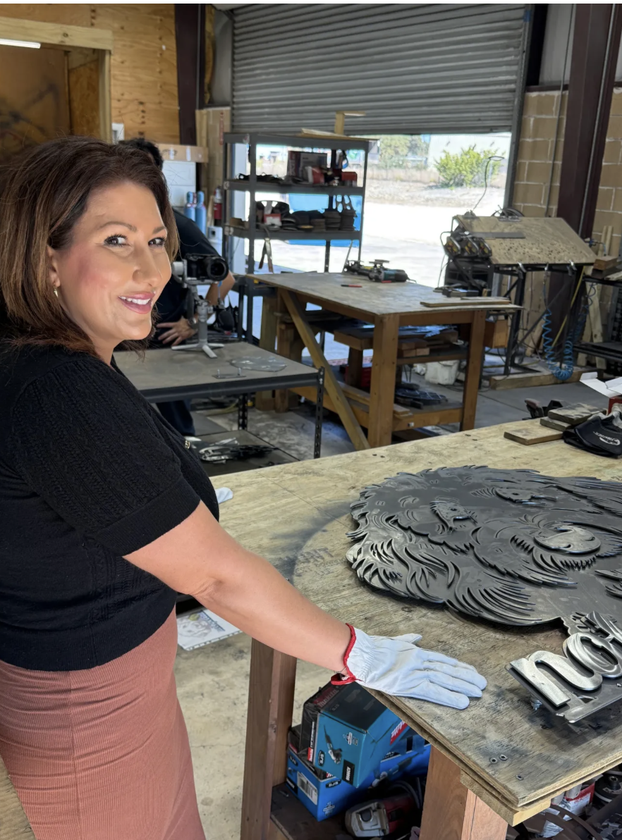 A woman with brown hair and a black top is standing in a workshop, smiling while working on a large metal decorative piece shaped like a lion's face. The workshop has wooden tables, shelves with tools and materials, and a large open garage door letti