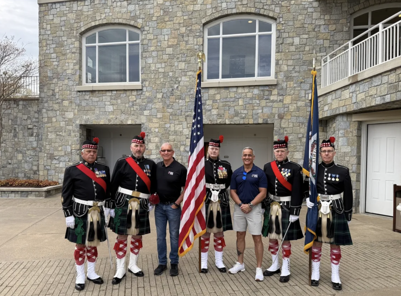 Seven men standing in front of a stone building, four dressed in traditional Scottish military uniforms with kilts, and three in casual clothing, holding flags, during daytime.