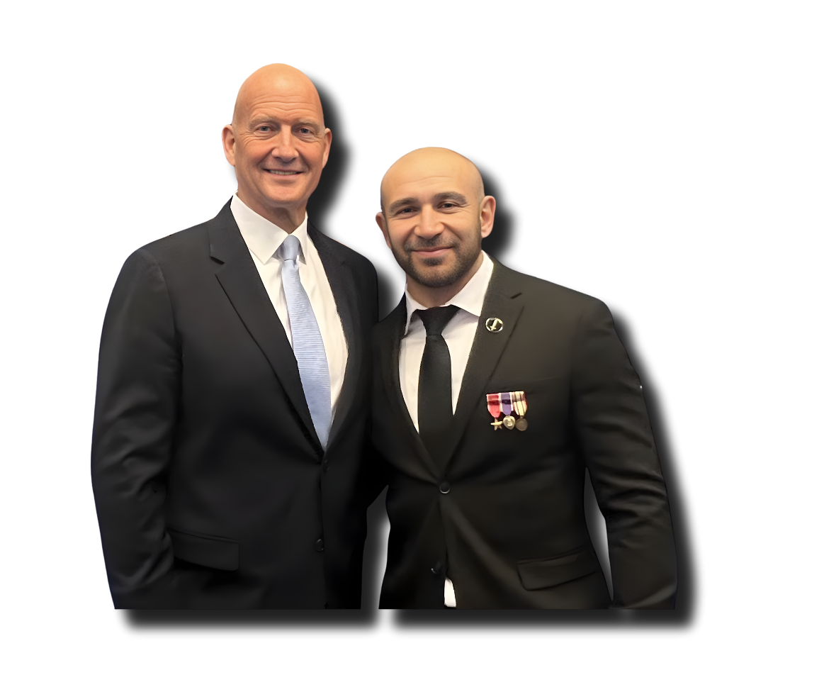 Two men in formal suits standing close together, smiling, against a black background. The man on the left is tall, with a bald head, wearing a dark suit, white shirt, and light blue tie. The man on the right is shorter, with a beard, wearing a dark suit, white shirt, black tie, and medals on his chest.