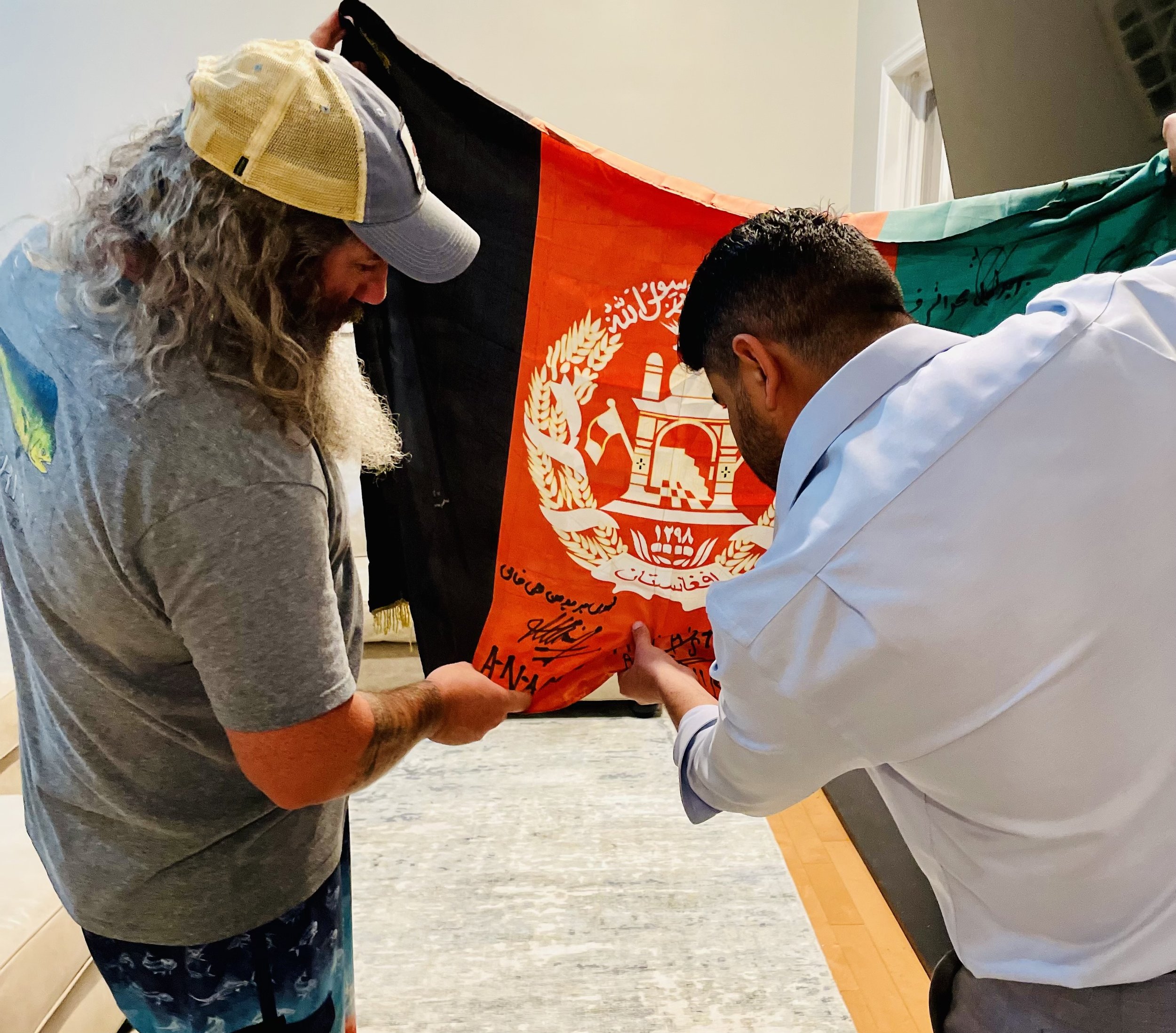 Two men participating in a formal handover or signing ceremony, holding an orange flag with Arabic and Afghan national symbols. One man has long, curly gray hair and a beard, wearing a baseball cap and a gray shirt. The other man, with short dark hai